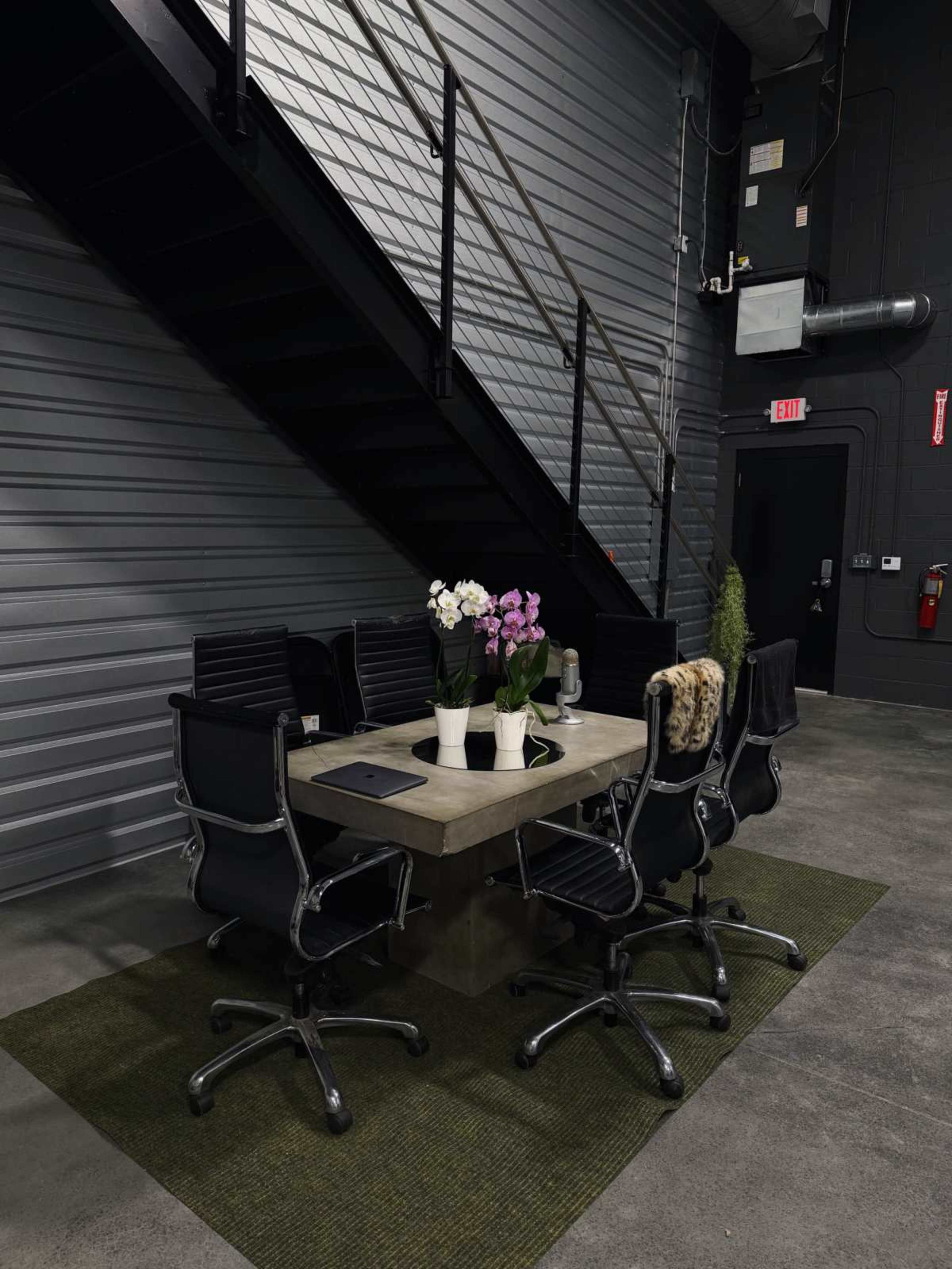 A conference table surrounded by black office chairs, with a decorative arrangement of flowers, is situated next to an industrial staircase against a gray wall.