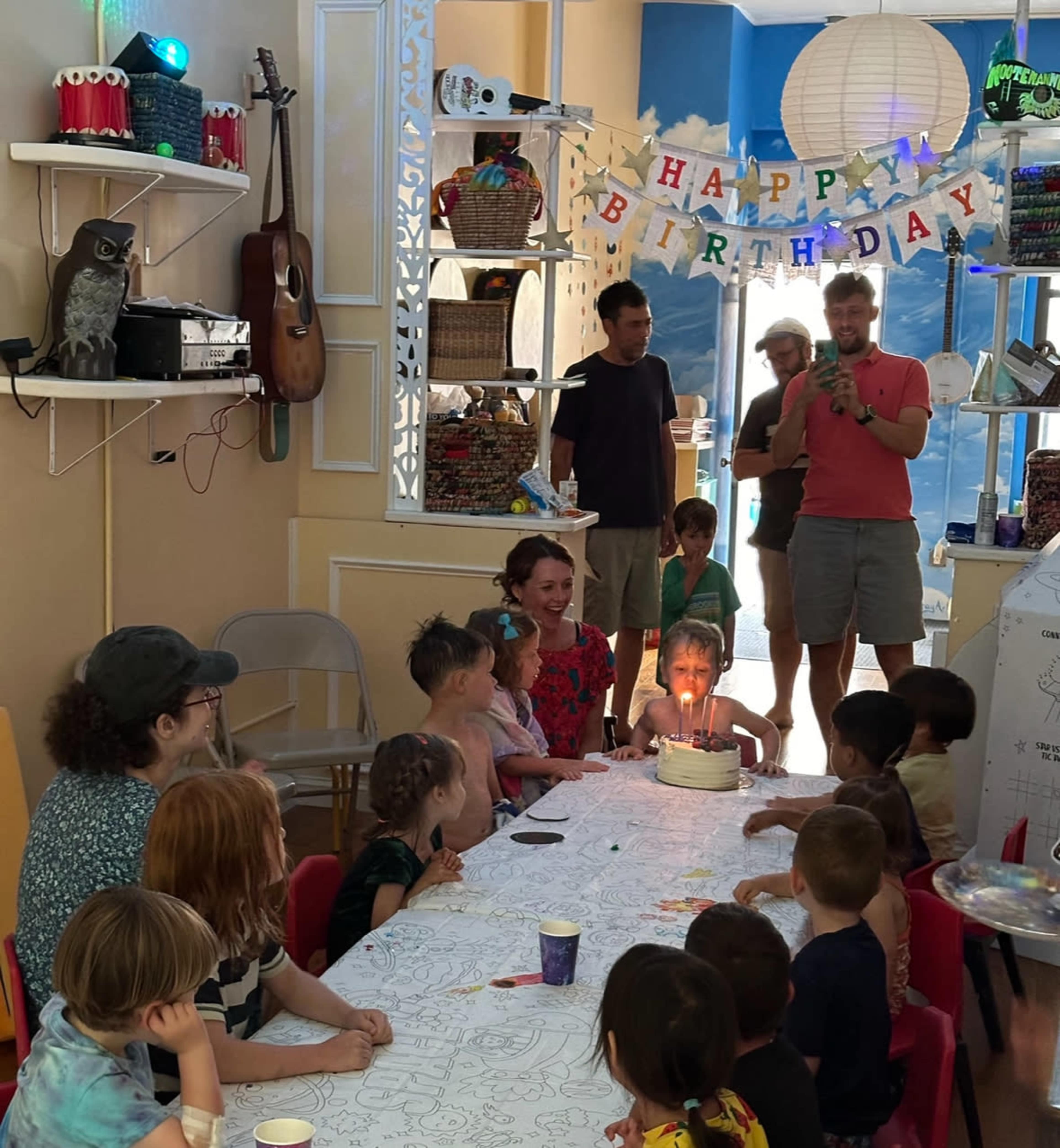 A birthday celebration is taking place in a colorful room with children seated around a long table, while an adult lights a cake surrounded by festive decorations.