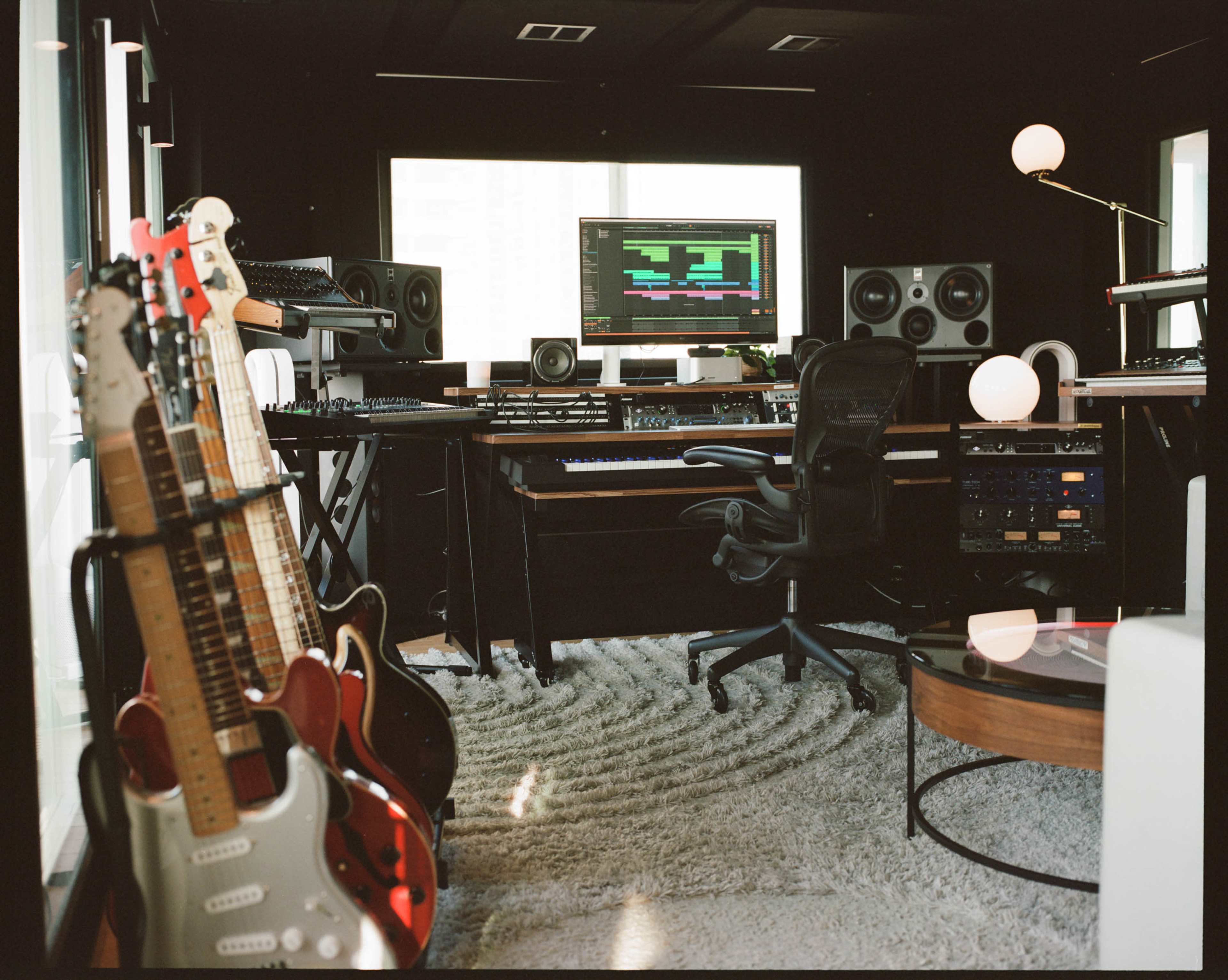 The image shows a modern music production studio featuring several guitars on stands, a central workstation with a computer and audio equipment, and large speakers in a sleek, well-lit space.