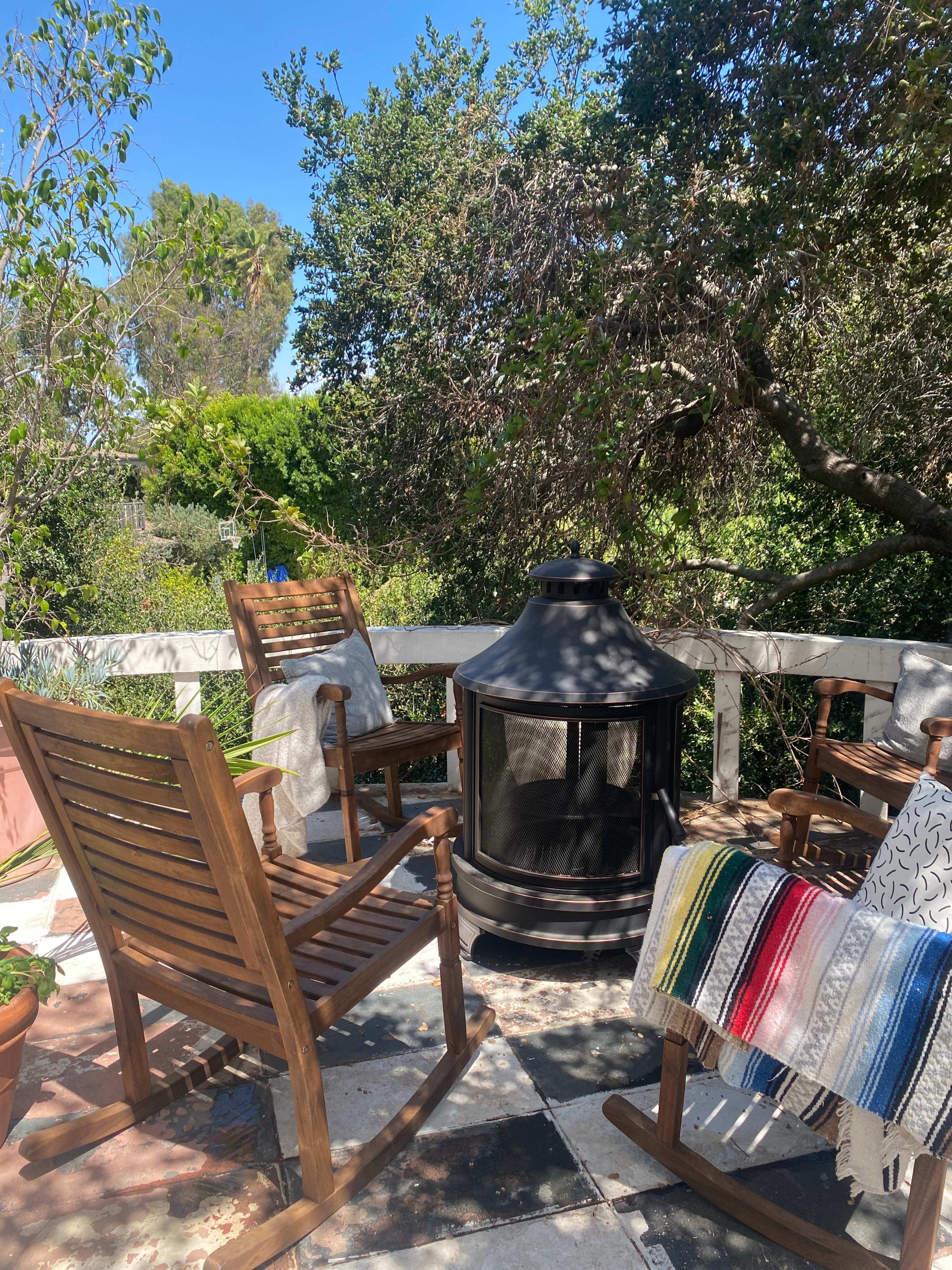 A seating area with wooden chairs and a black outdoor fireplace is surrounded by lush greenery.