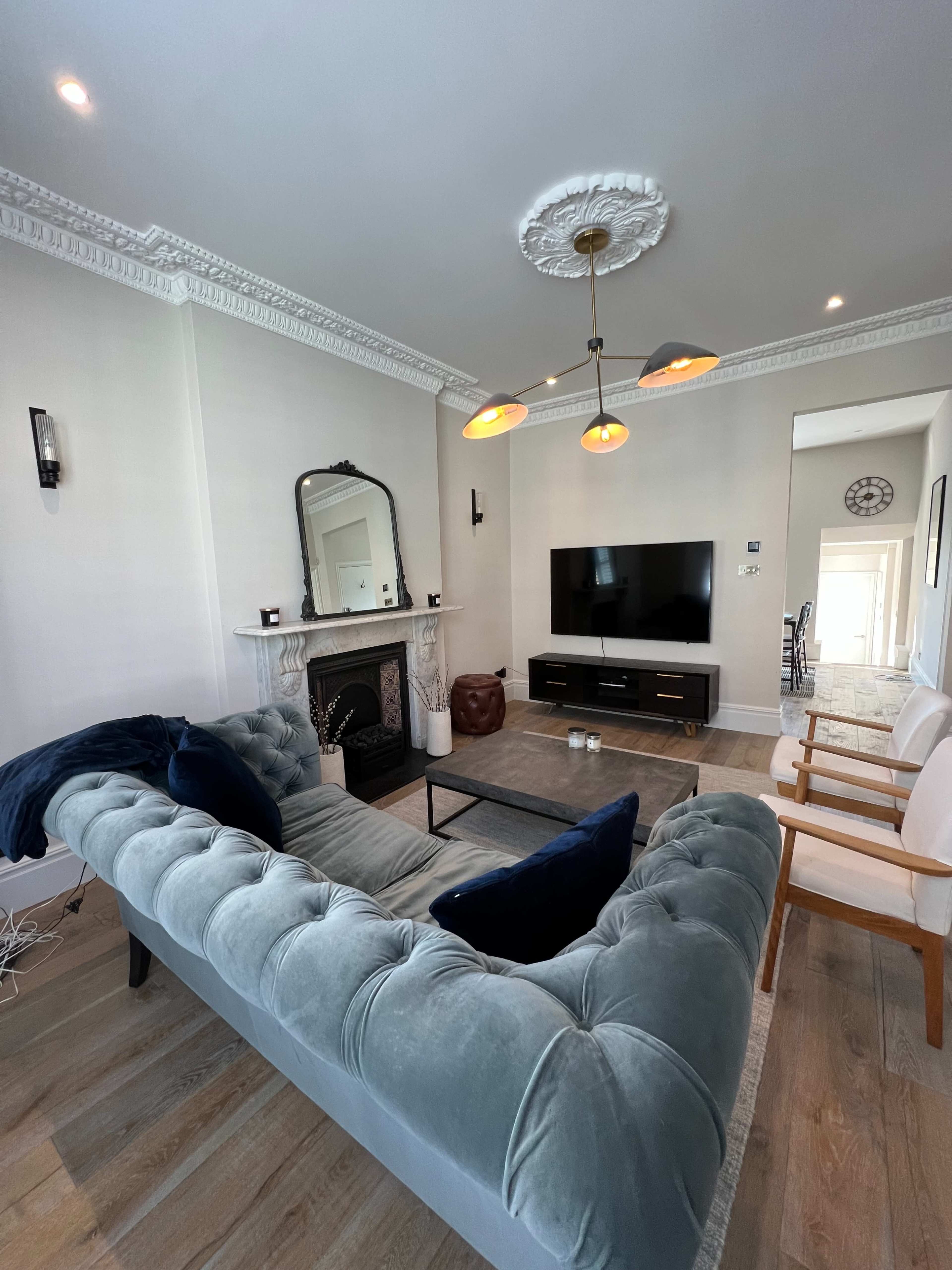 A stylish living room features a tufted sofa, modern light fixtures, and a wall-mounted television opposite a fireplace.