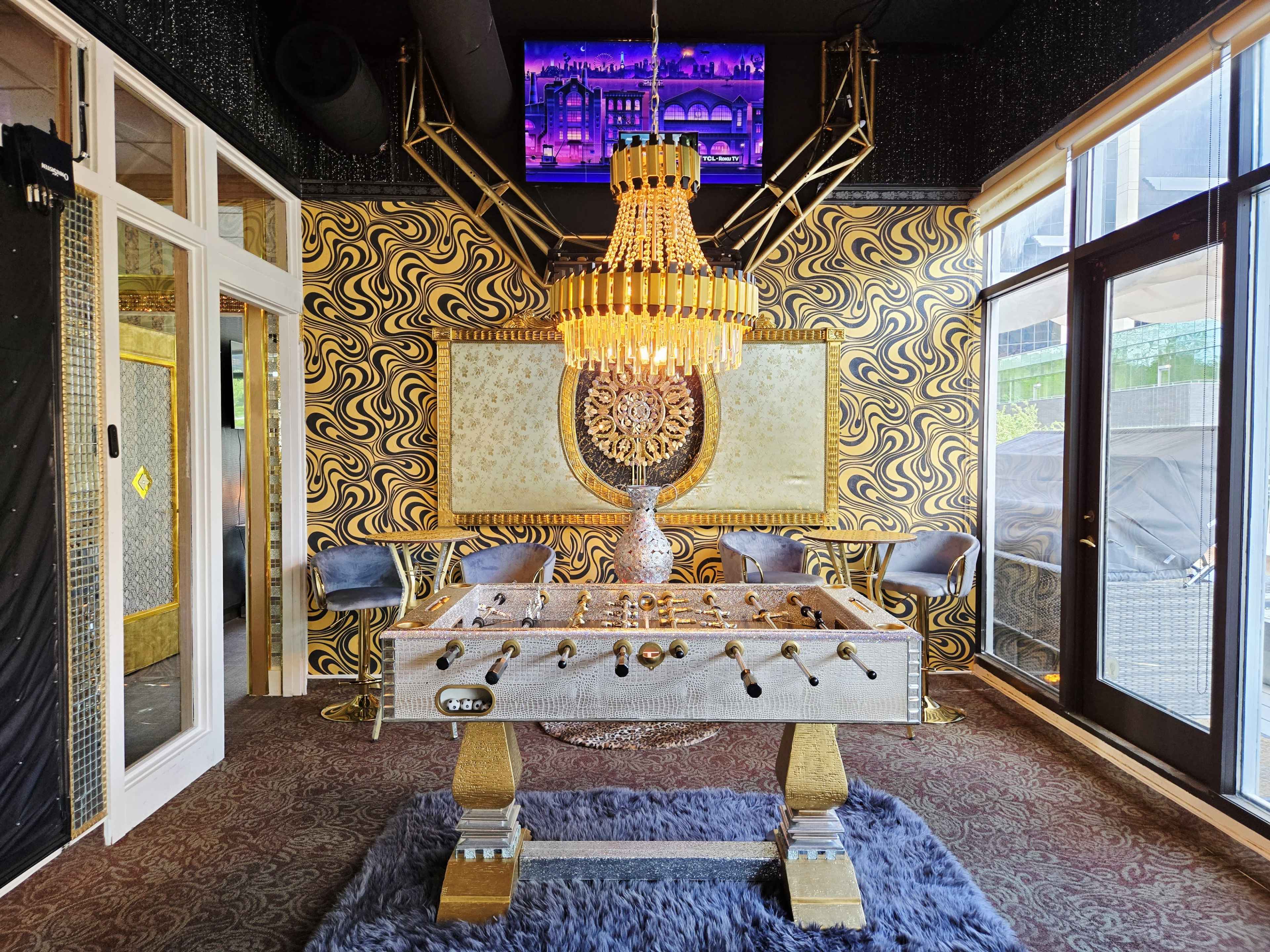 The scene features a stylish game room with a foosball table, a chandelier, and decorative walls in a vibrant pattern.