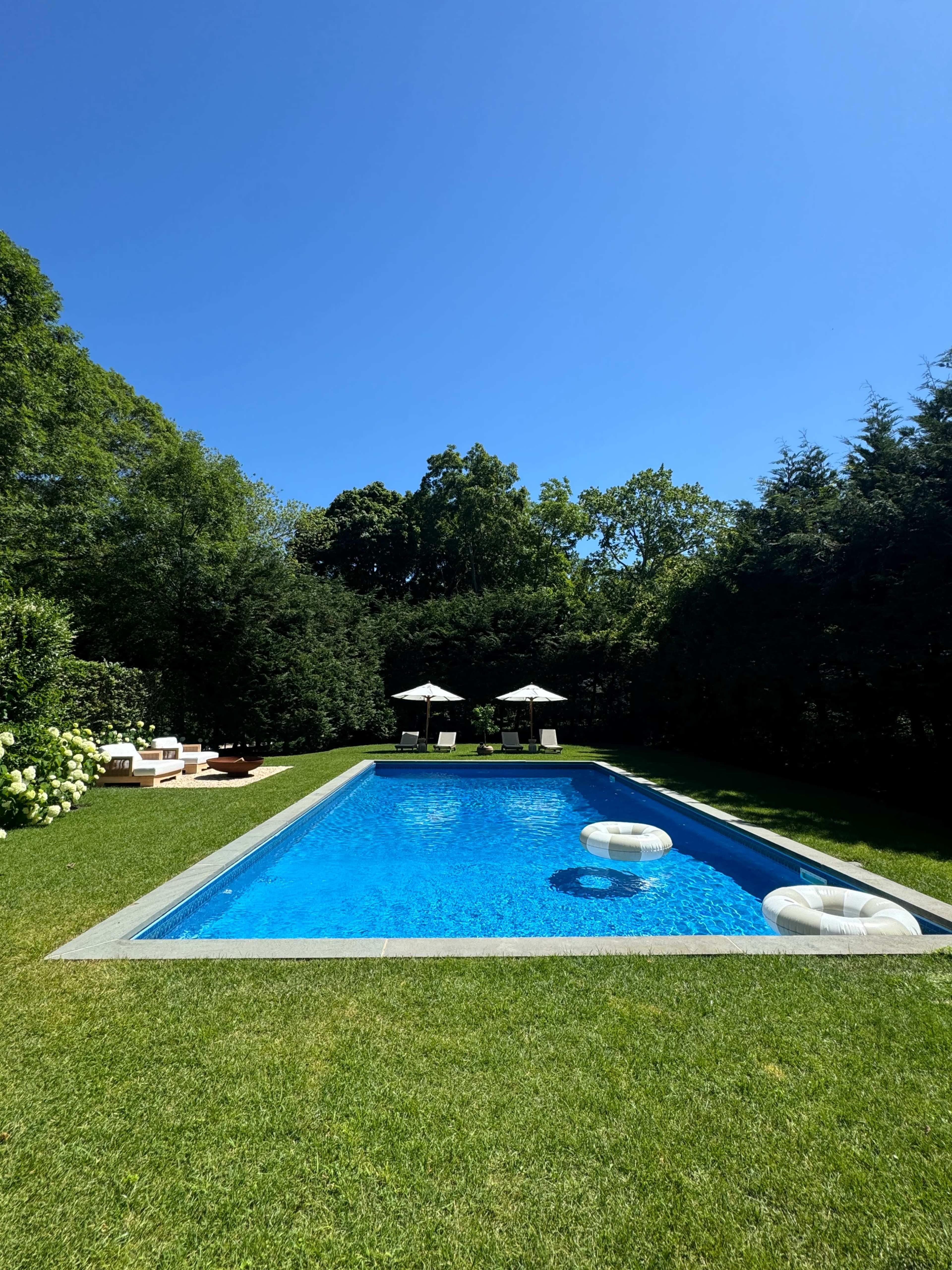 A rectangular swimming pool with two floating rings is surrounded by green grass and trees, featuring two lounge chairs beneath two umbrellas at one end.