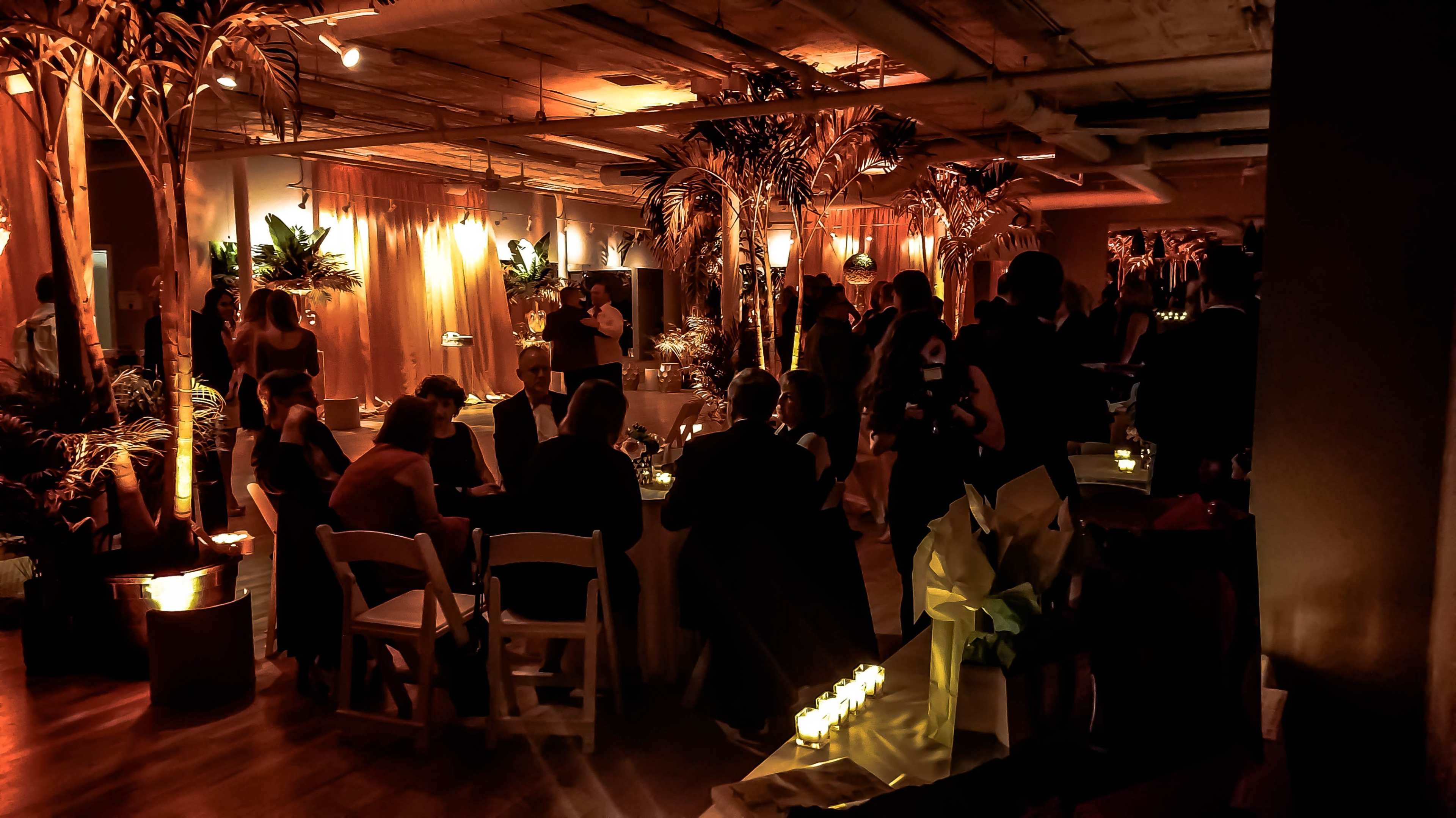 A dimly lit event space is decorated with tropical plants and candles, with guests seated at tables and mingling.