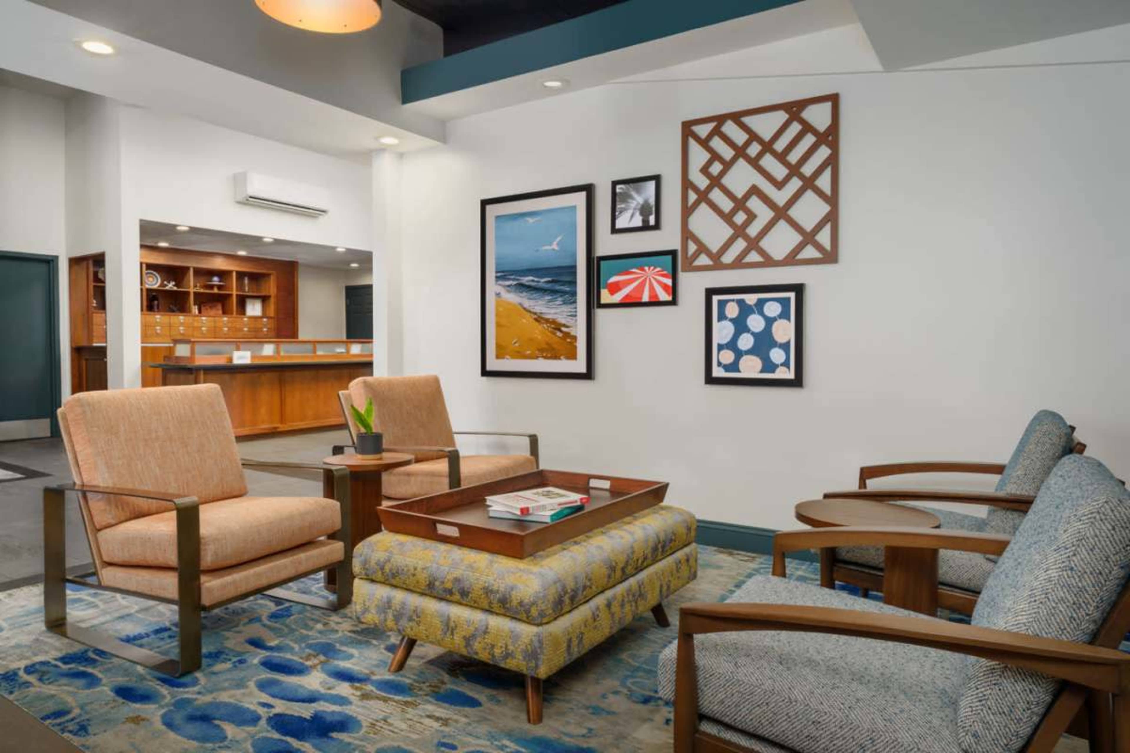 The image shows a modern lobby seating area featuring two armchairs, a patterned ottoman, and a blue area rug, alongside framed artwork on the wall.