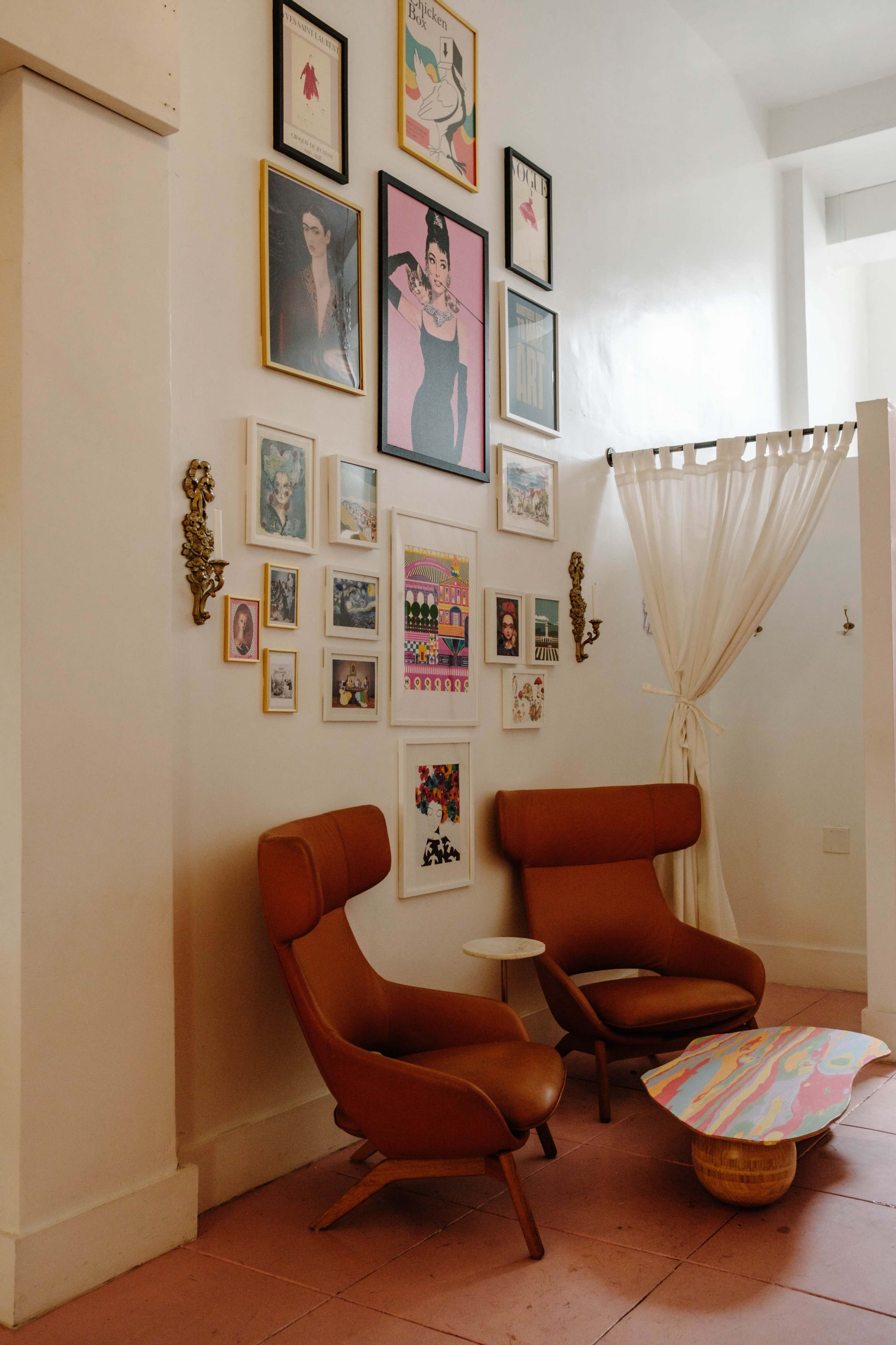 The image shows a corner of a room with two brown lounge chairs, a small white table, and a wall adorned with an assortment of framed artworks and photographs.