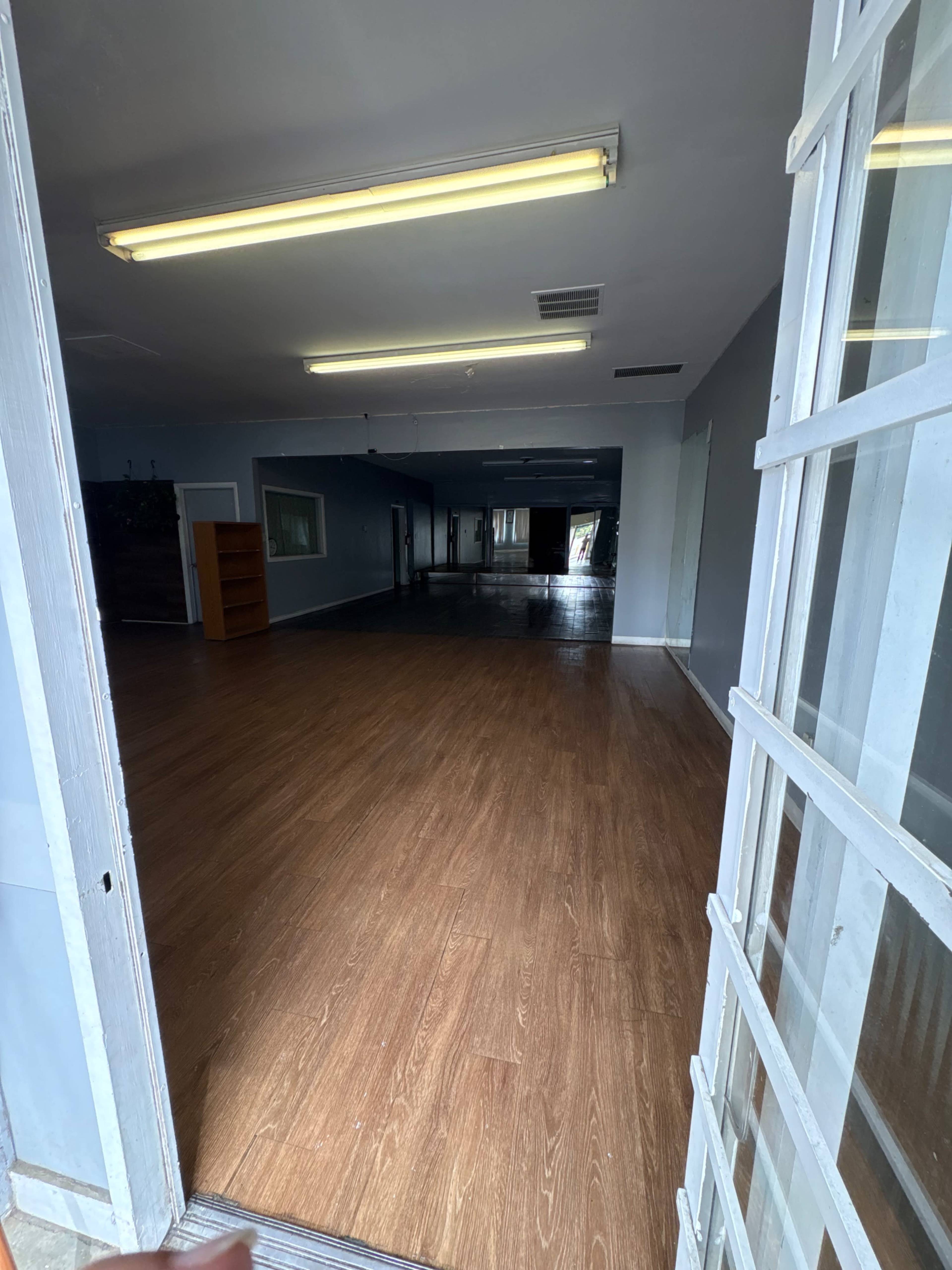 The image shows an empty interior space with a wooden floor, visible lighting fixtures, and an open door leading outside.