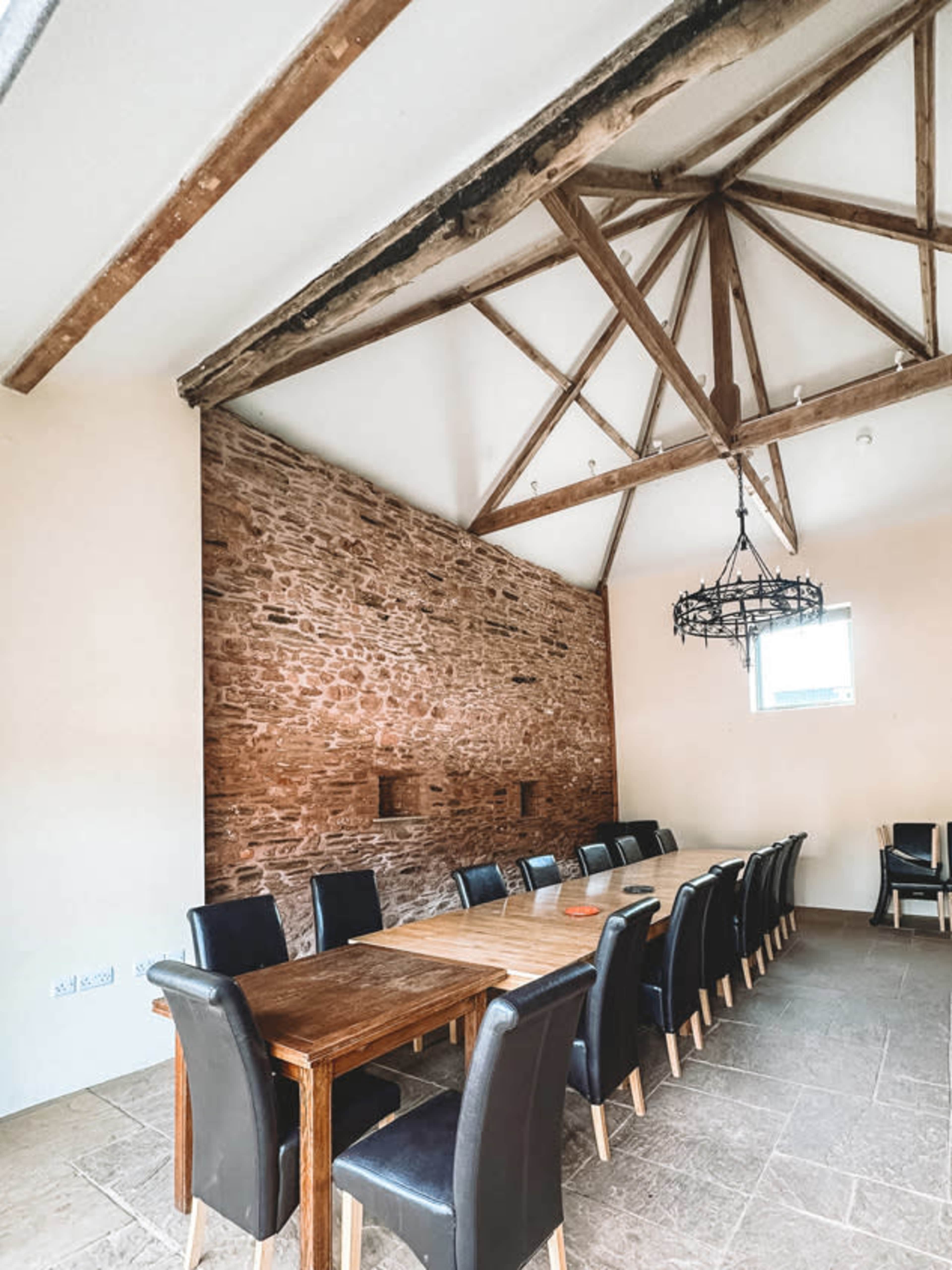 A spacious dining area featuring a long wooden table surrounded by black upholstered chairs, with a stone wall and exposed wooden beams above.