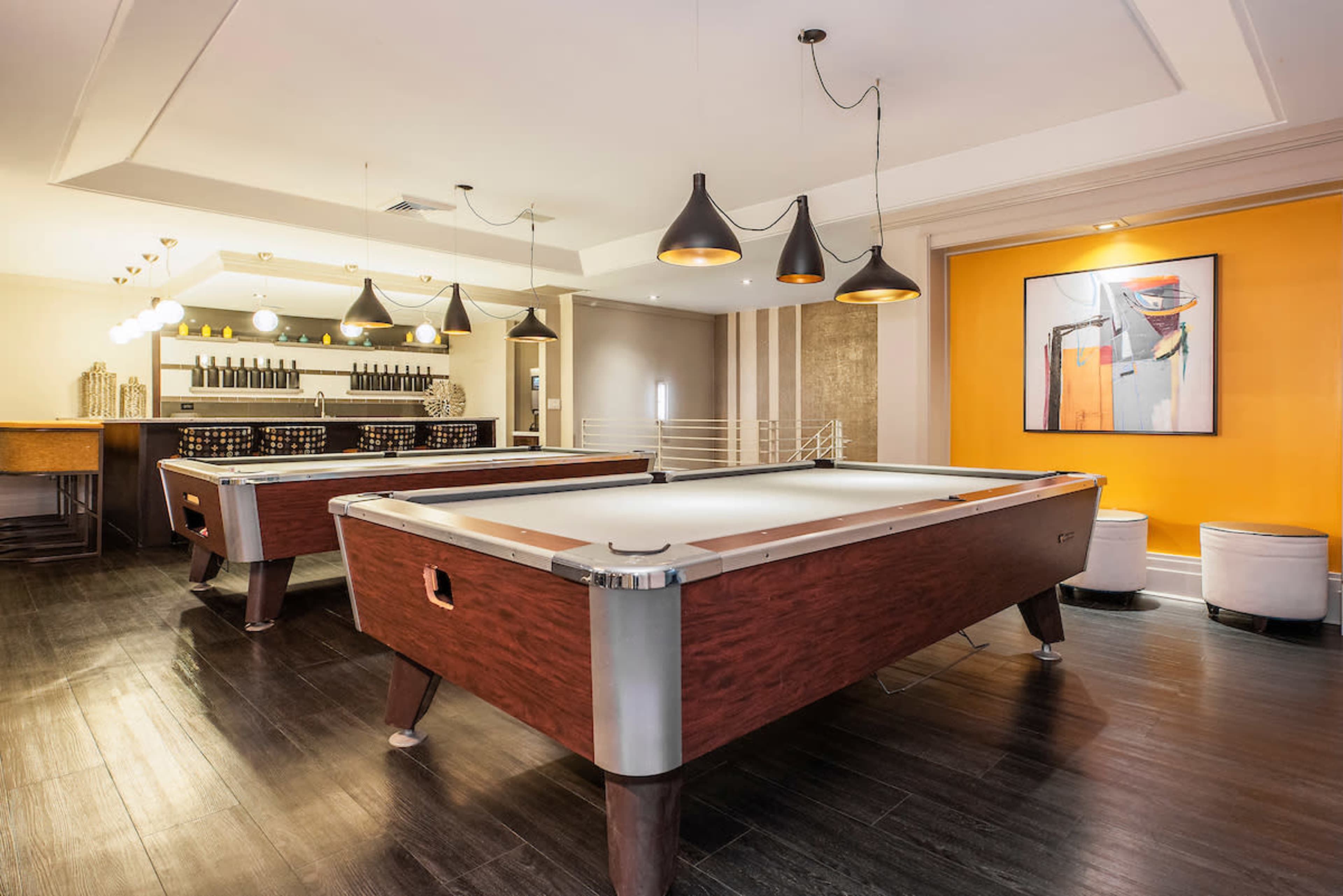 A modern game room featuring two pool tables, a bar area with bottles, and stylish pendant lights.
