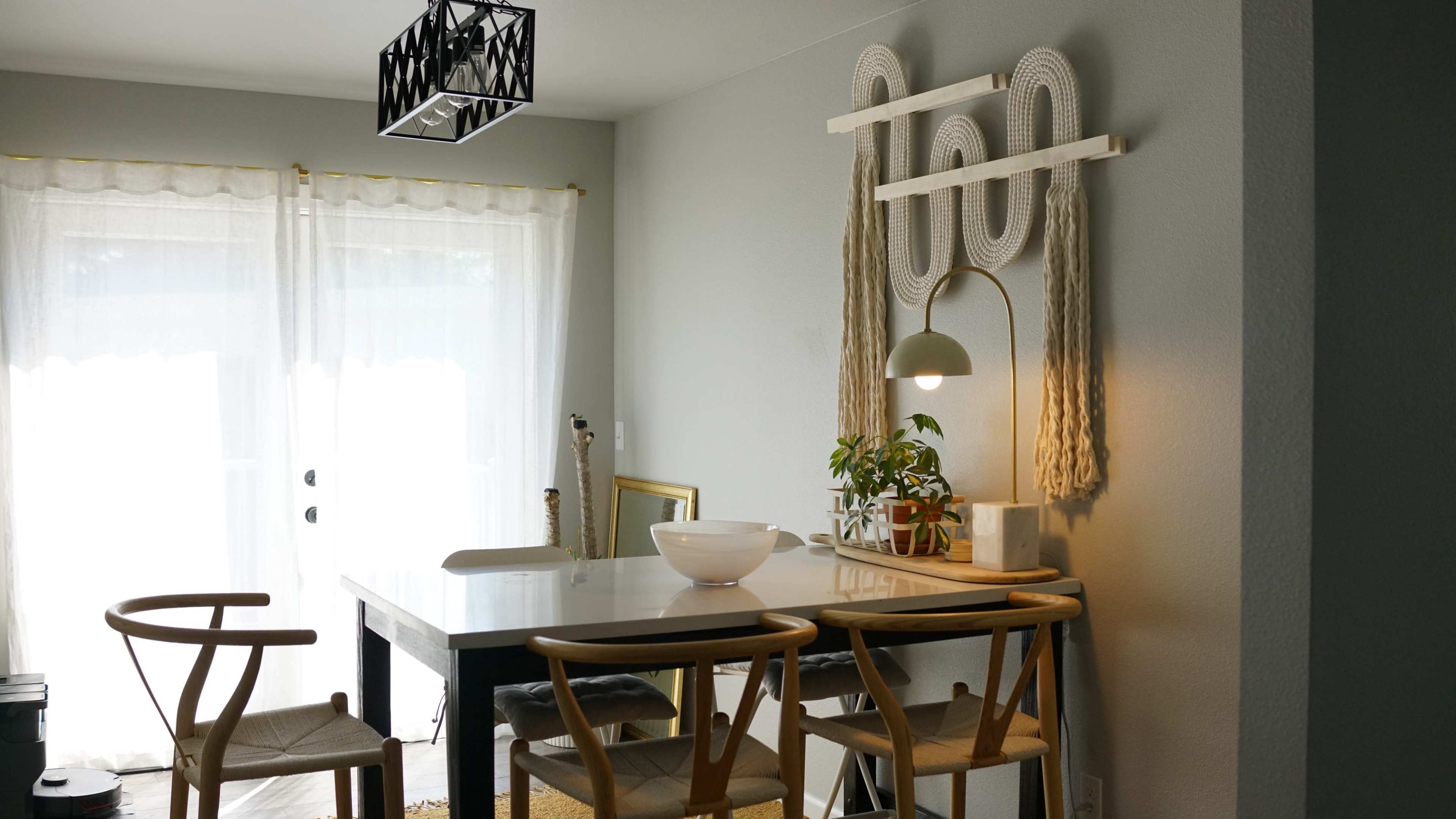 A minimalist dining area features a small table with three chairs, a plant, and decorative wall art, illuminated by a pendant light.