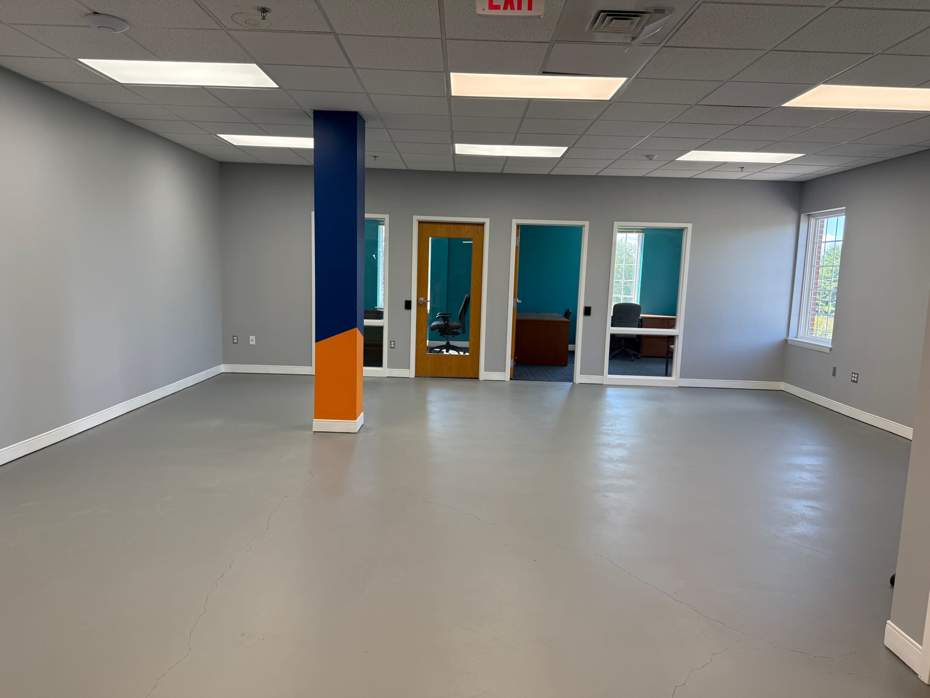 An empty office space features grey floors, blue and orange accent walls, and multiple glass-paneled rooms.
