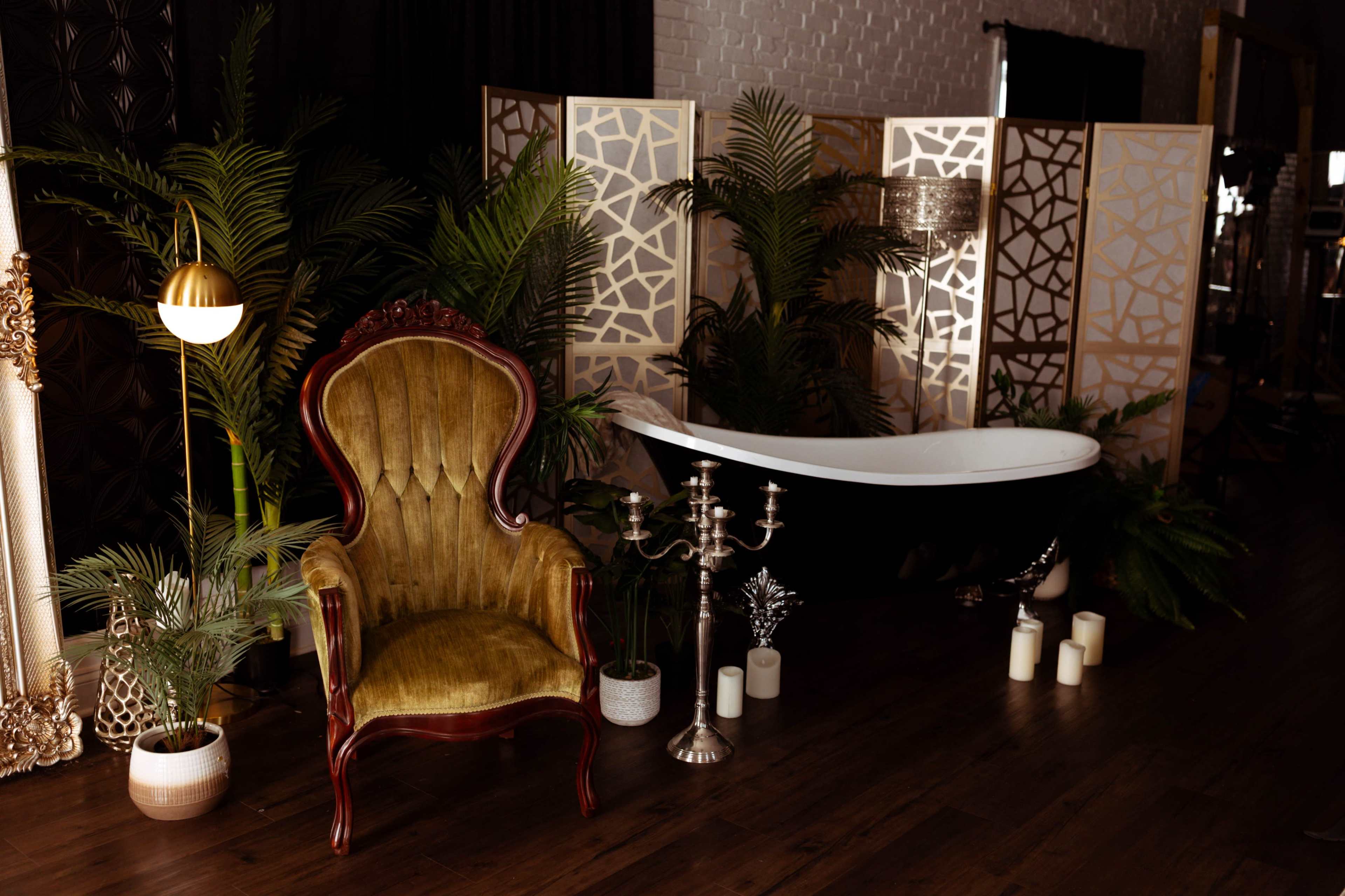 The scene features an elegant vintage chair beside a modern freestanding bathtub, surrounded by decorative screens, potted plants, and candles.