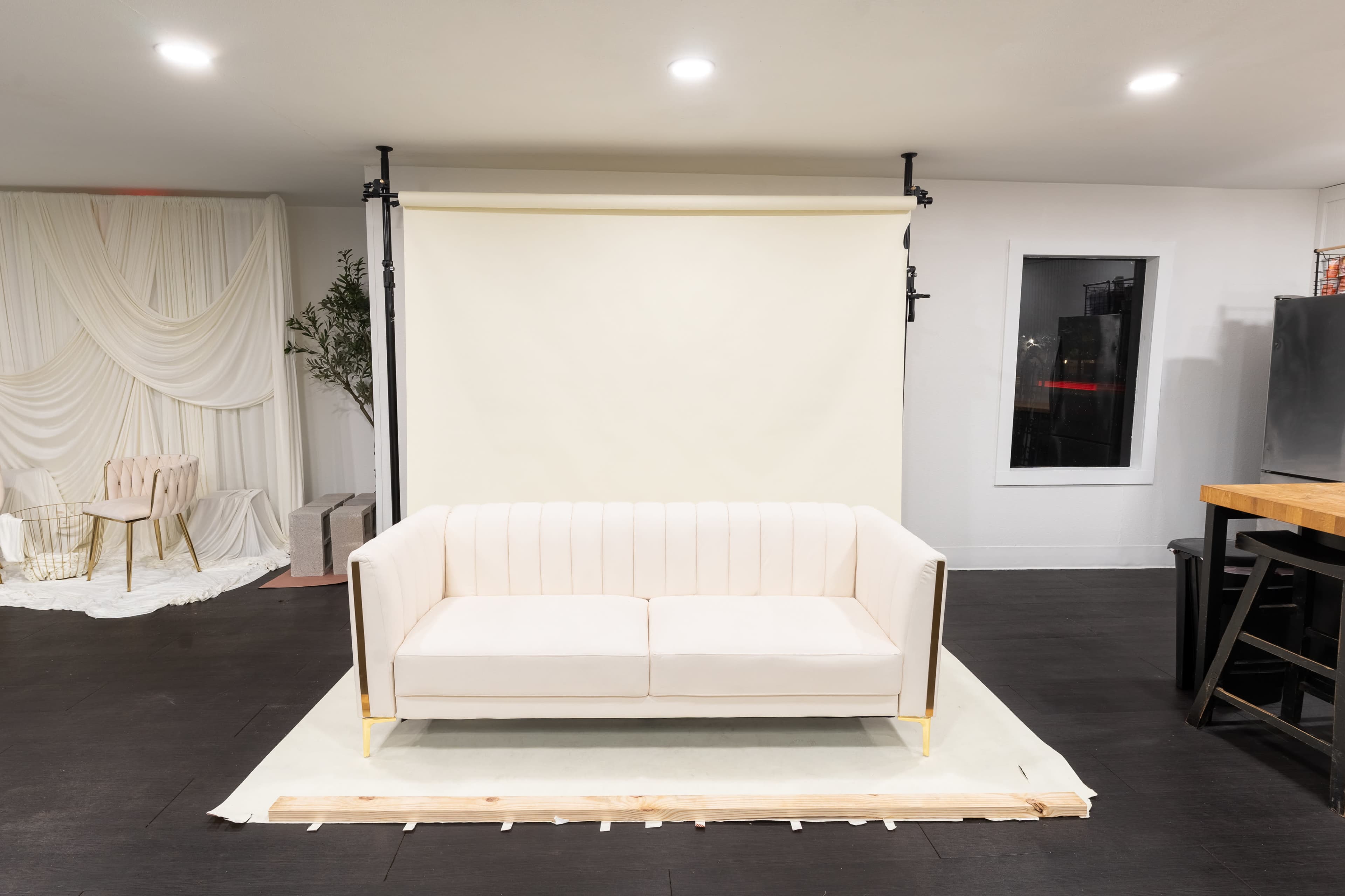 A modern, cream-colored couch is placed in front of a plain light-colored backdrop in a minimalist studio space.