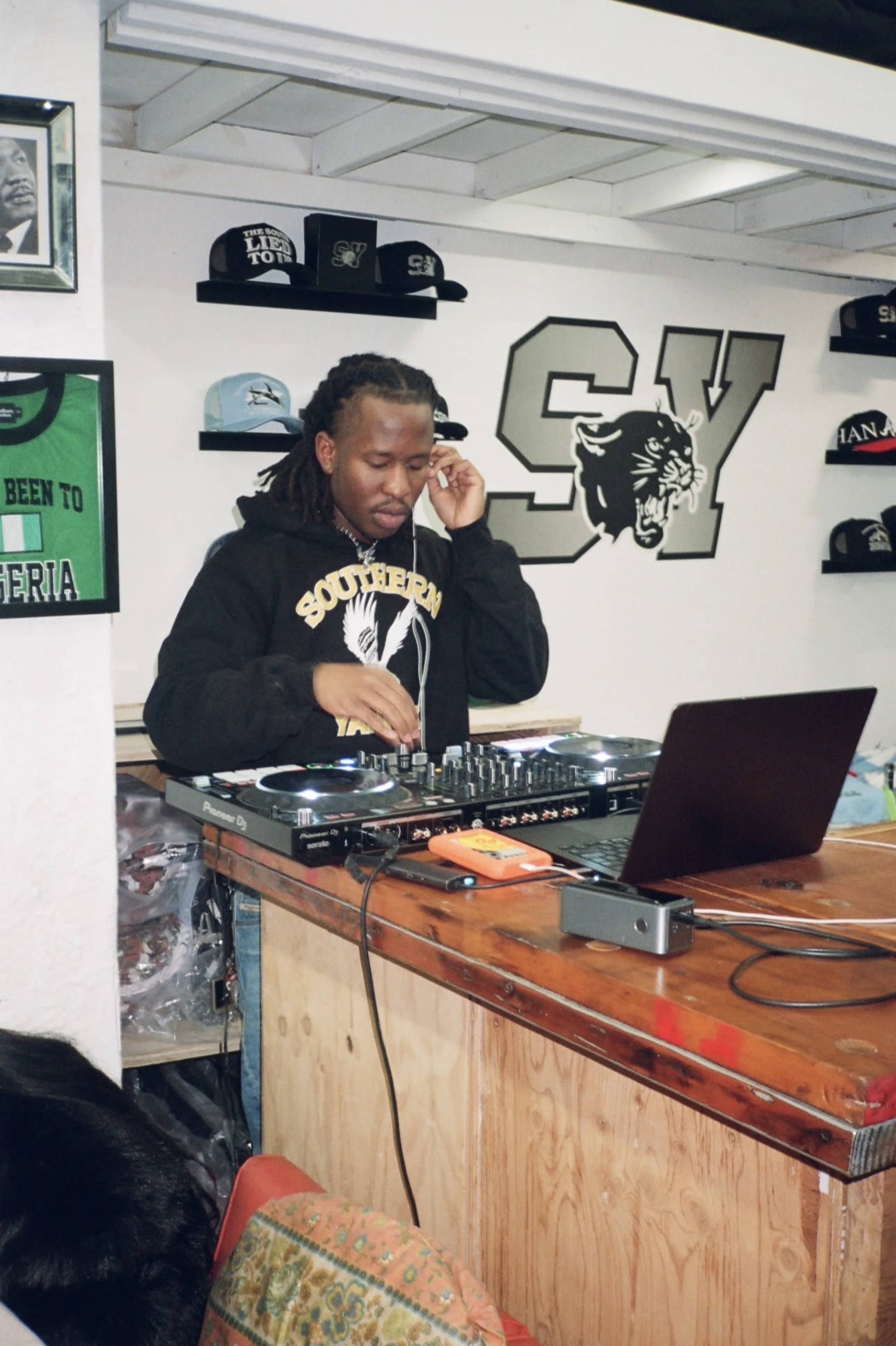 A DJ operates a mixing console in a shop adorned with various hats and apparel.