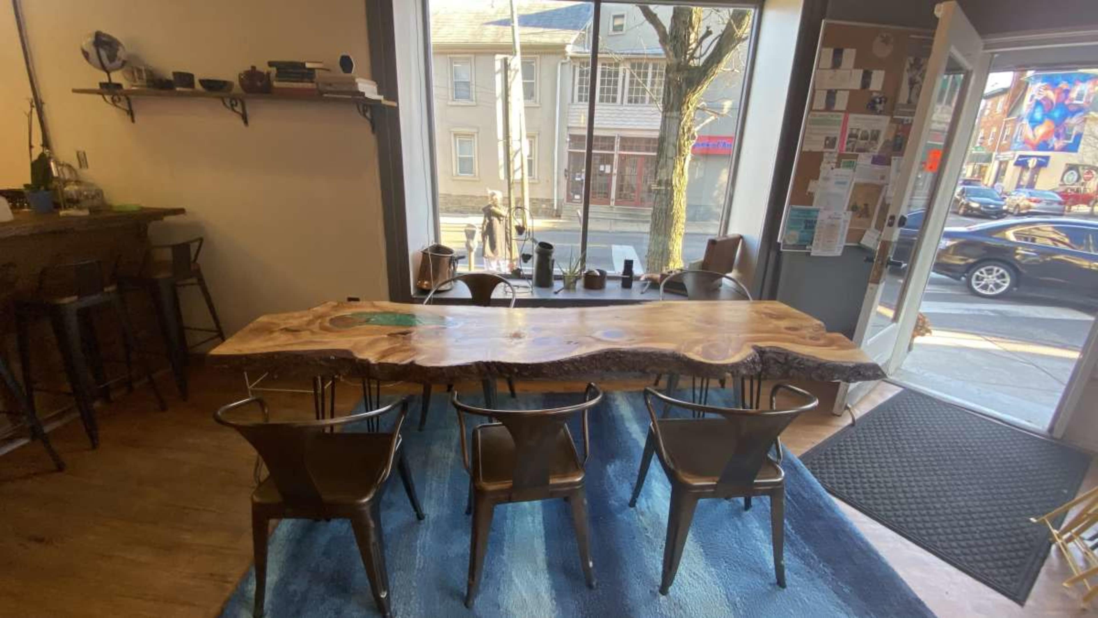A large wooden table with a live edge is surrounded by metal chairs in a well-lit room featuring a view of a street outside.