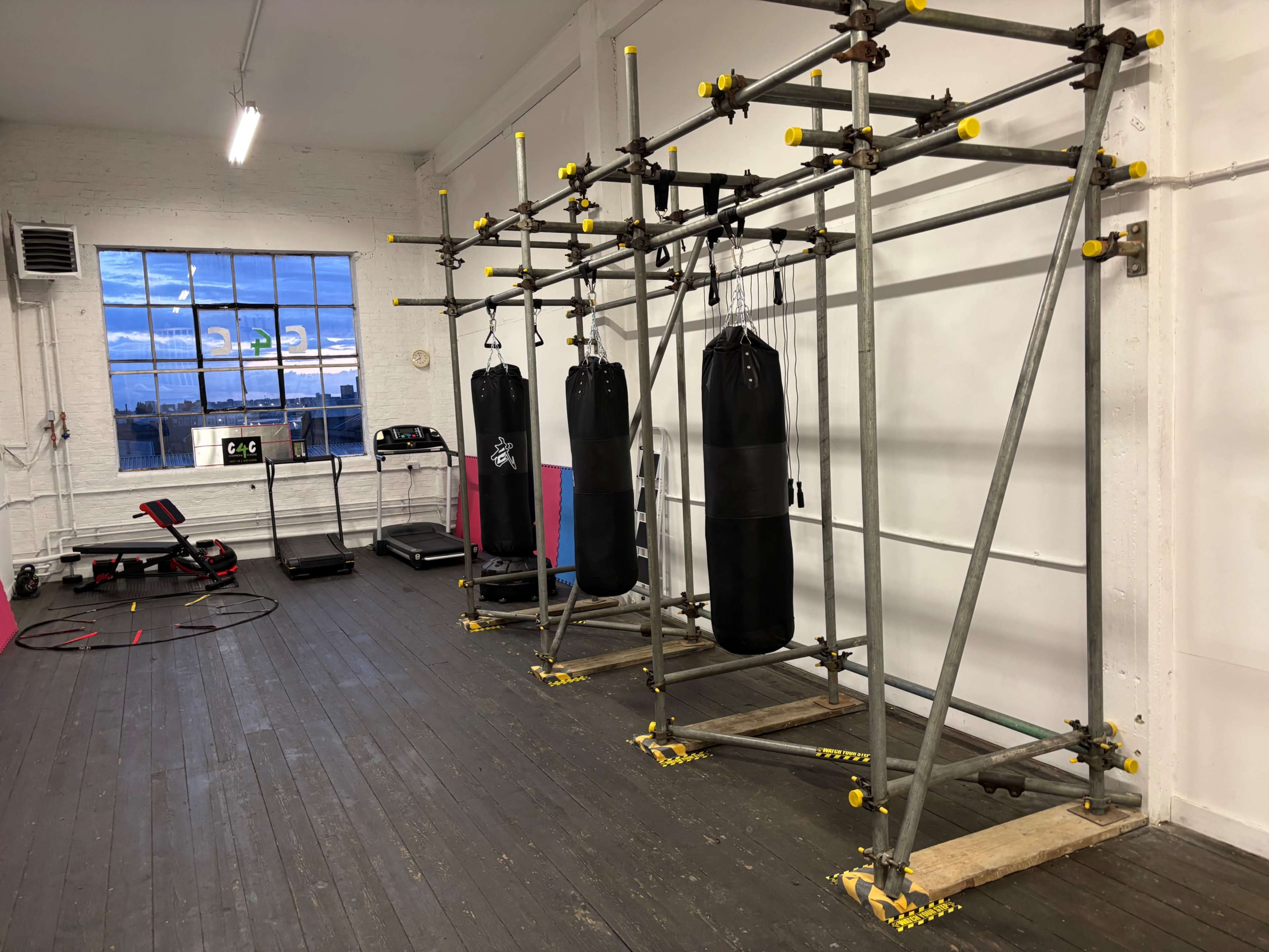 The image shows a gym interior with three boxing bags hanging from a metal frame, along with exercise equipment and large windows in the background.