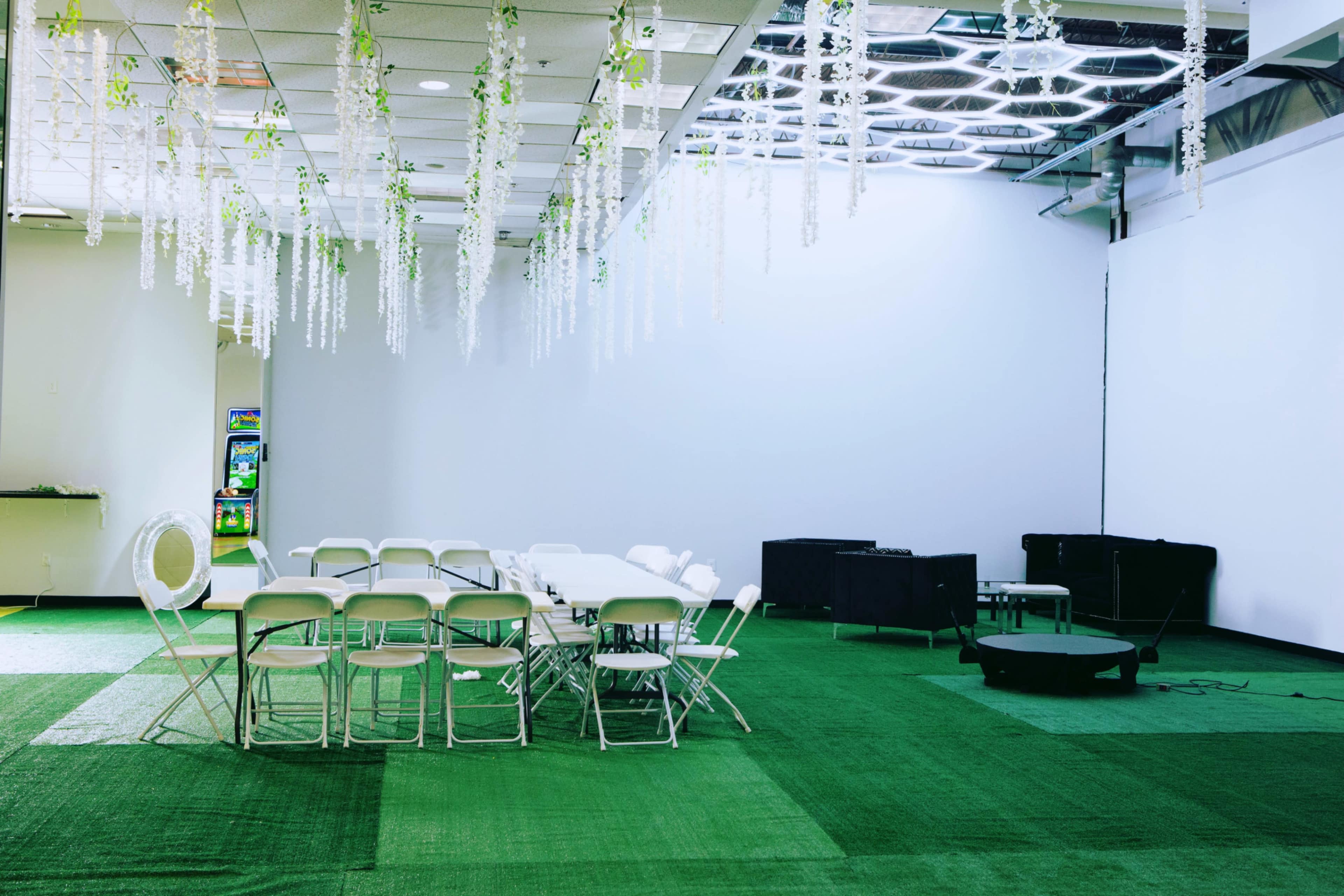 A room with green carpet features a circular table arrangement with white folding chairs and decorative hanging plants.