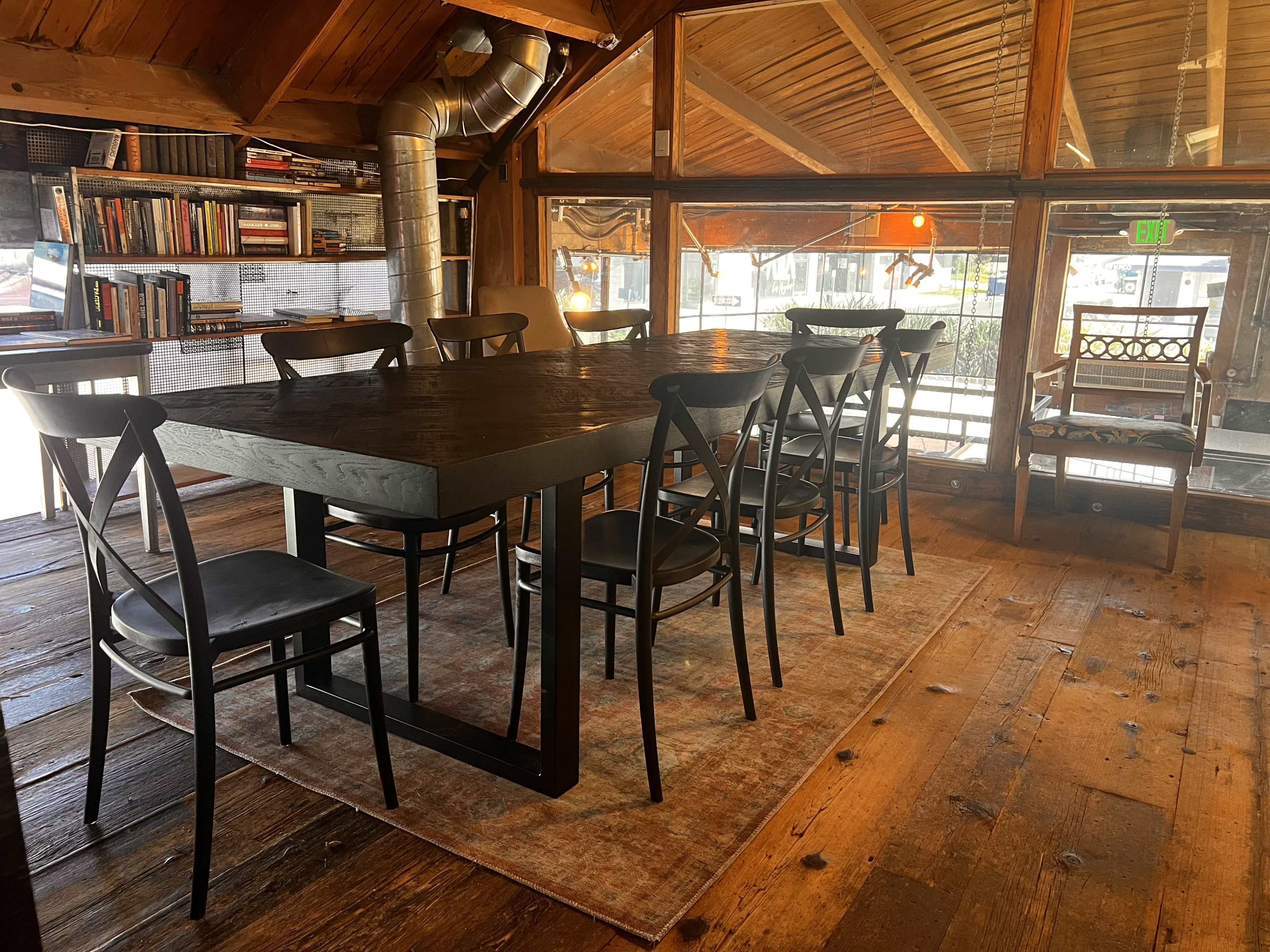 A large, black wooden table with eight chairs is set in a wooden-floored room featuring bookshelves and large windows.