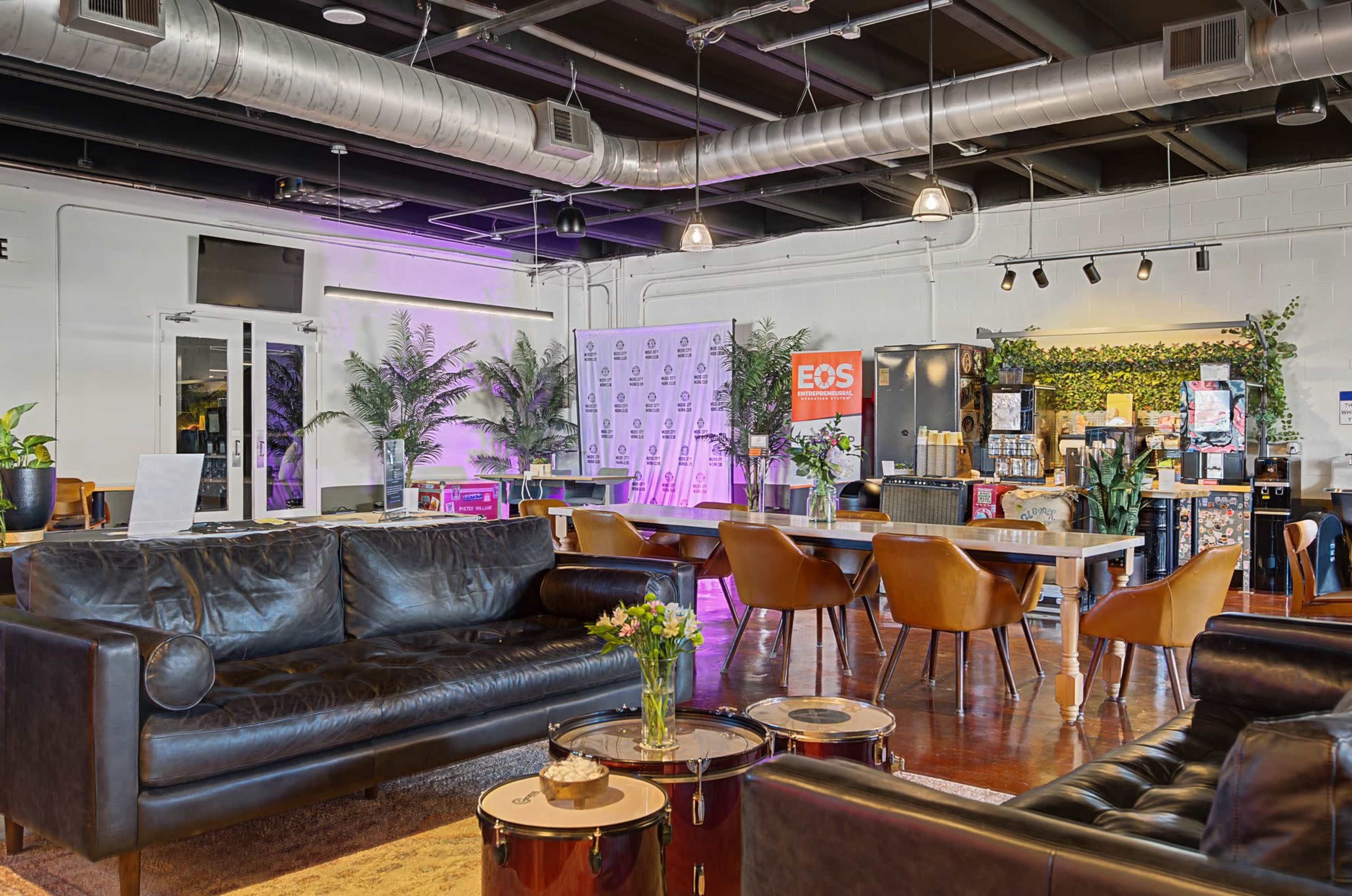 The image shows a modern, open interior space featuring dark leather sofas, wooden tables, and greenery, with a bar area in the background.