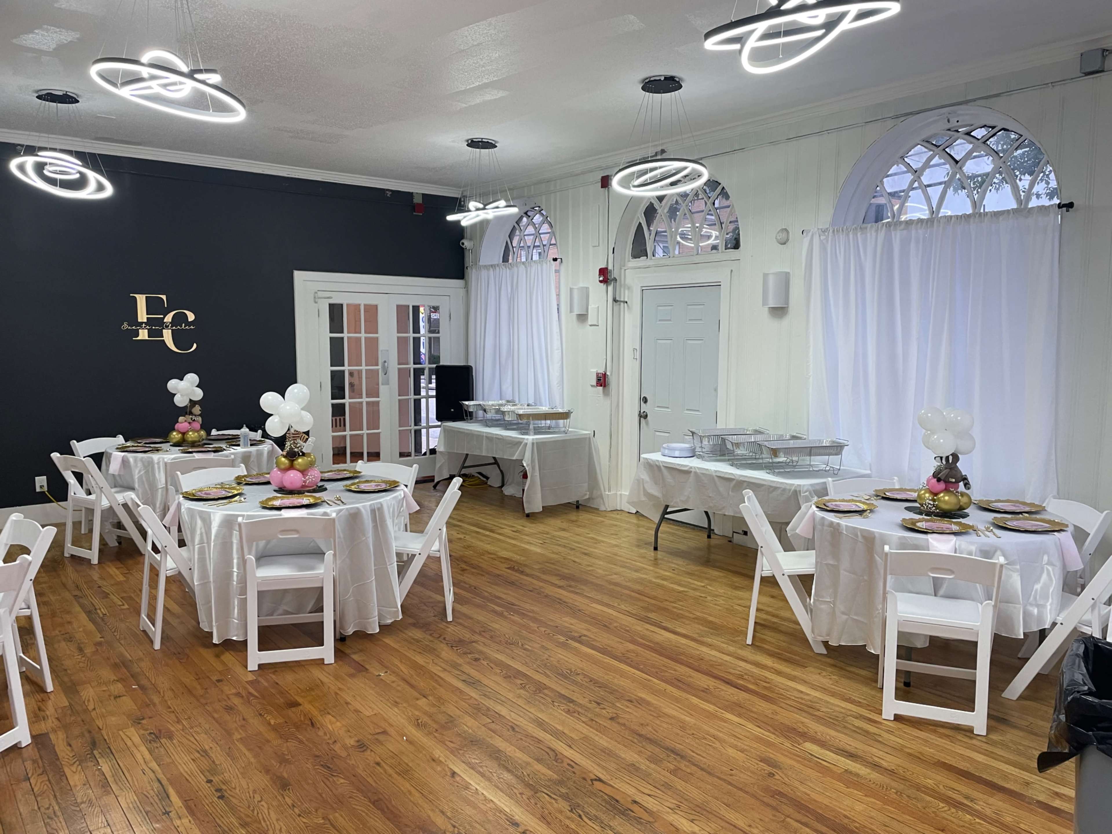 A well-lit event space with two round tables set for a meal, decorated with white tablecloths and arrangements of balloons, against a backdrop of a dark wall and large windows.