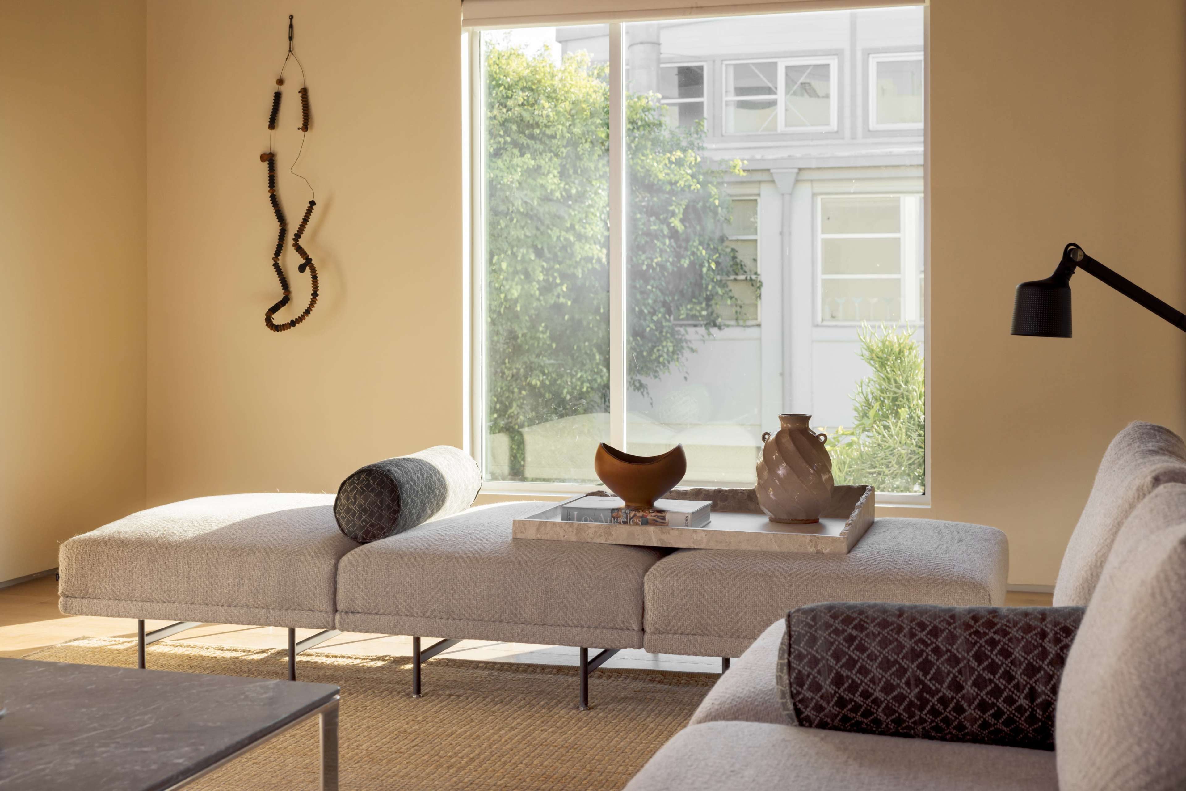 A modern living room features a light-colored sectional sofa, a coffee table with decorative items, and a large window looking out to a garden.