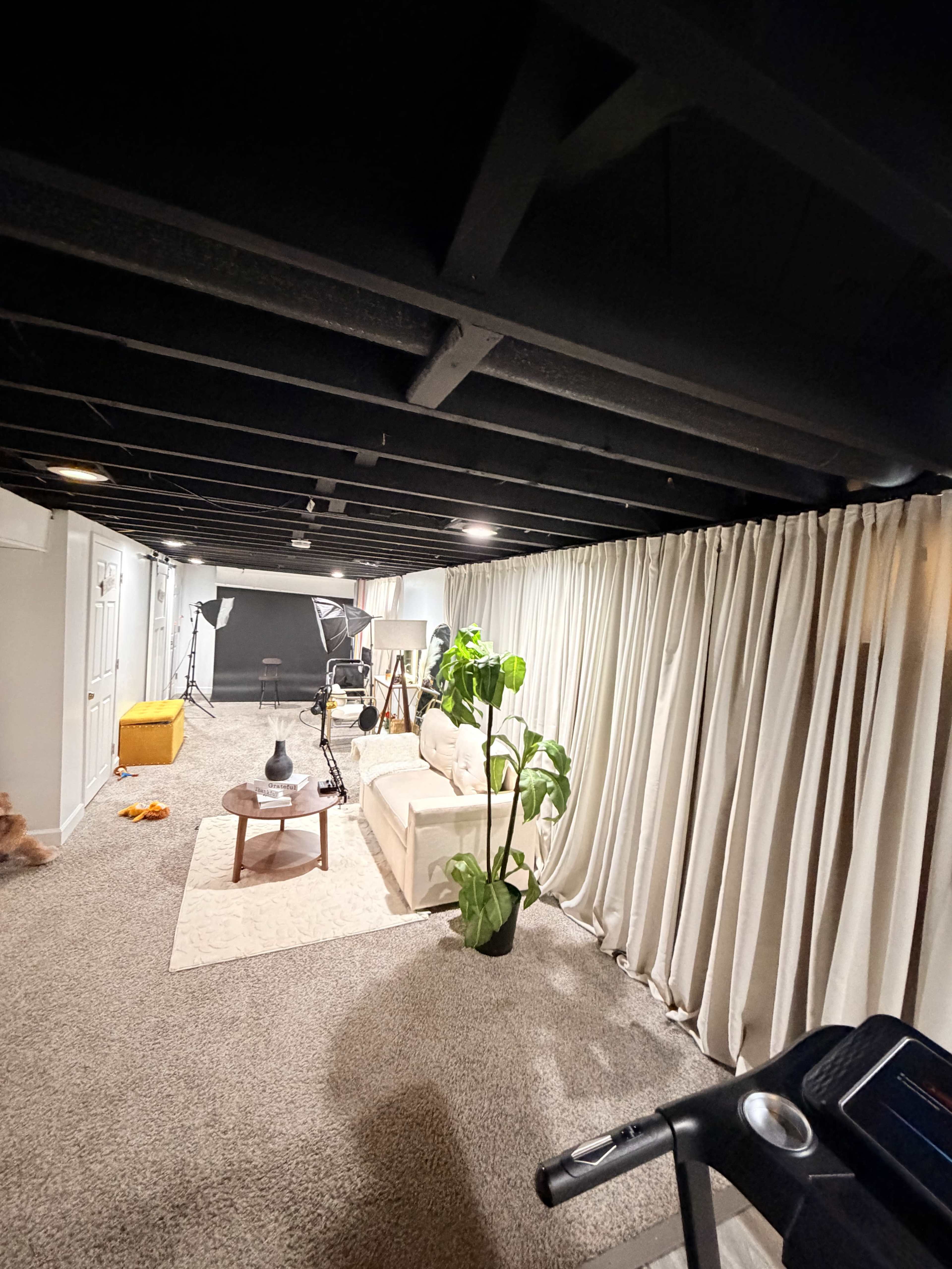 The image shows a basement room featuring a treadmill, a couch, potted plant, and curtains, with studio lighting equipment in the background.
