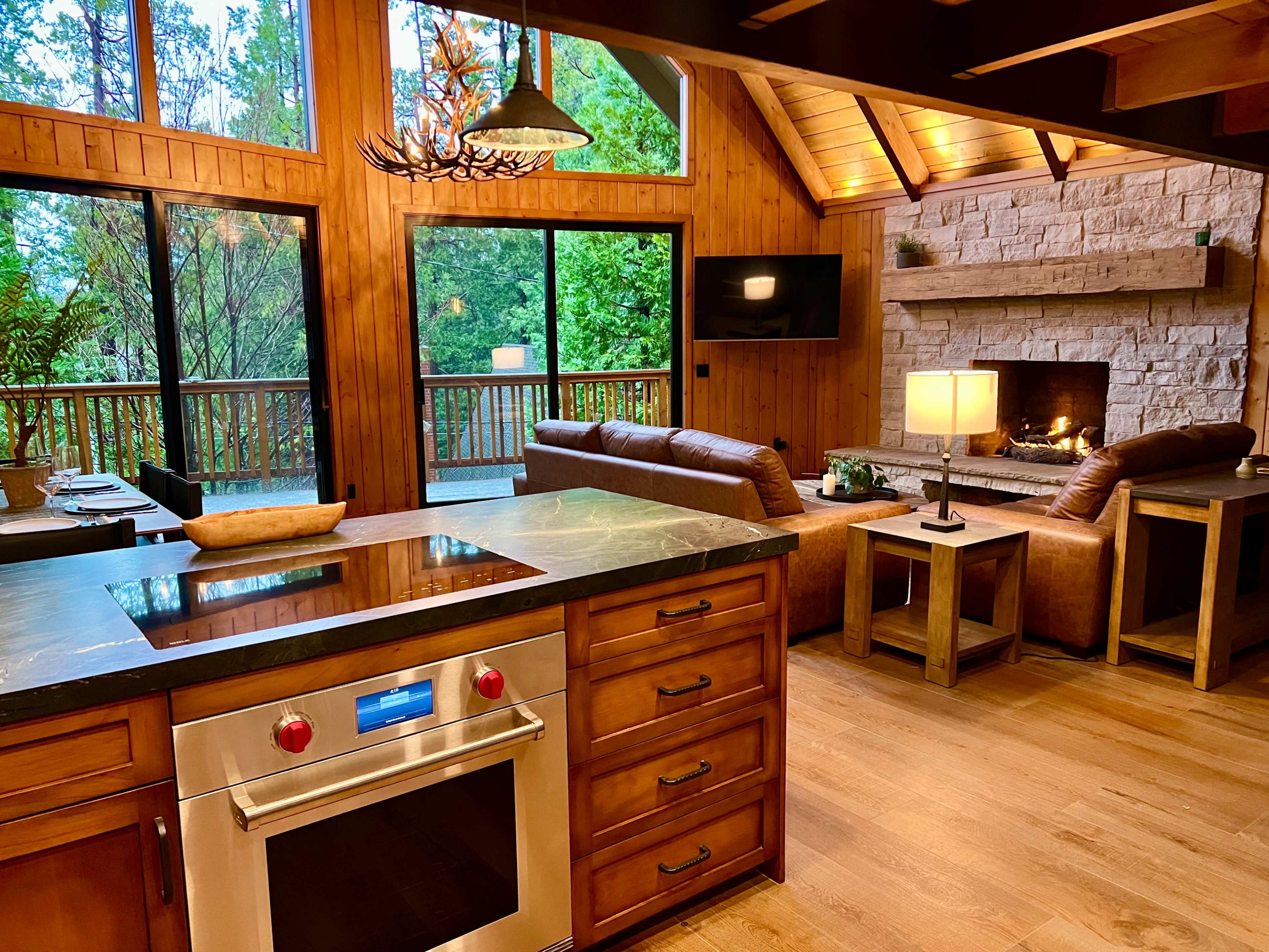 A modern kitchen and living area with wooden walls, a stone fireplace, large windows, and a cozy seating arrangement.