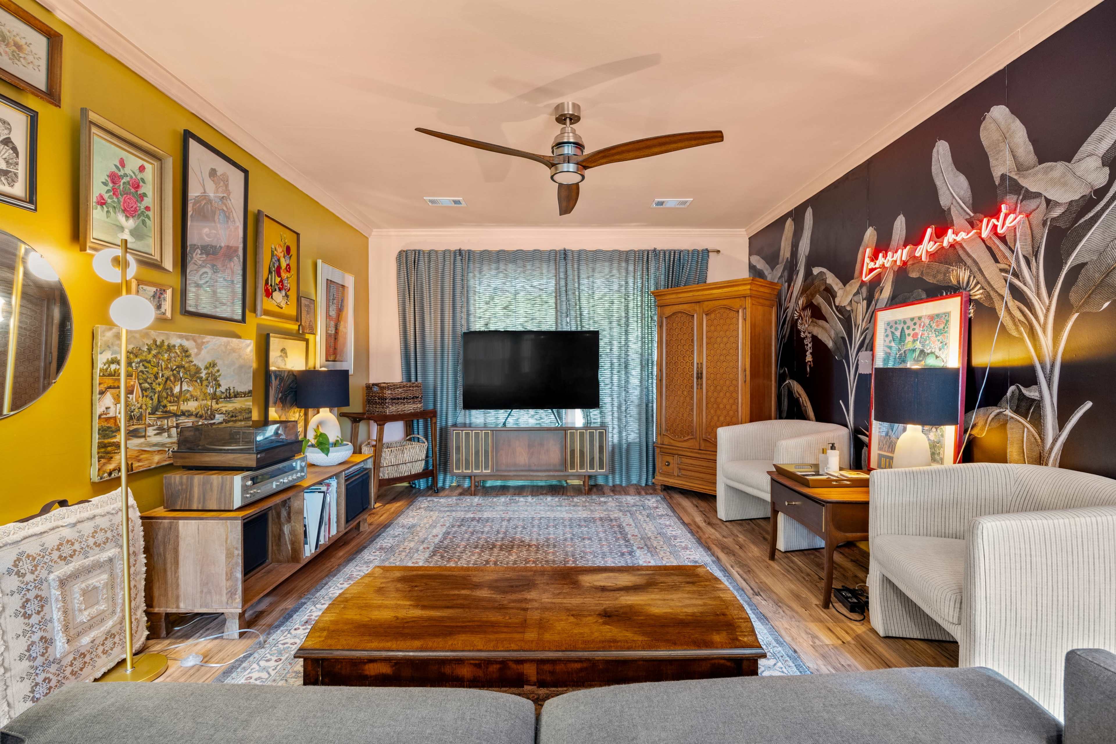 The living room features a television mounted on a wall, surrounded by artwork, a wooden coffee table, two white armchairs, and a decorative rug.