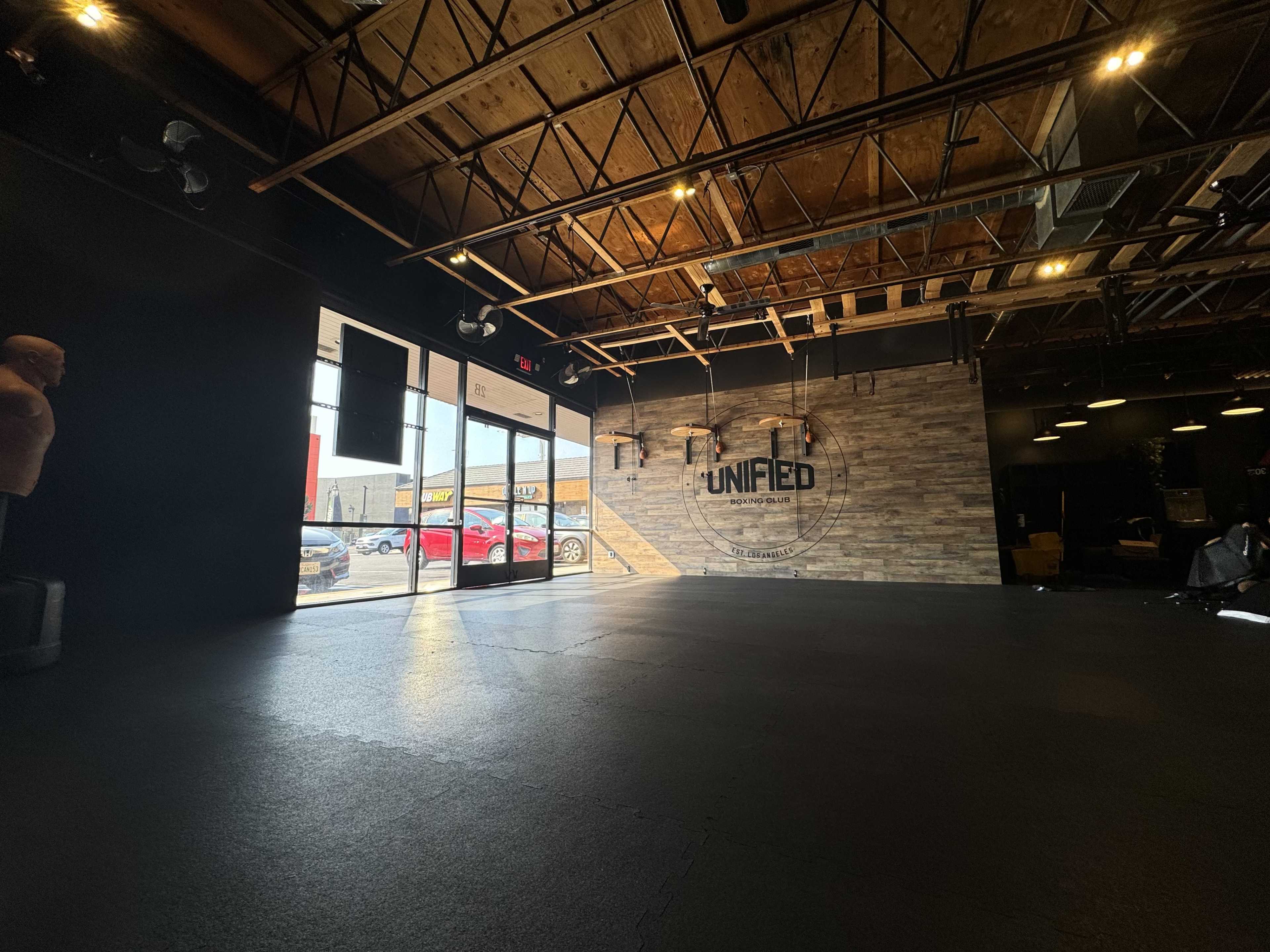 The image shows an empty gym interior with a black rubber floor, exposed wooden beams on the ceiling, and a logo on a textured wall.