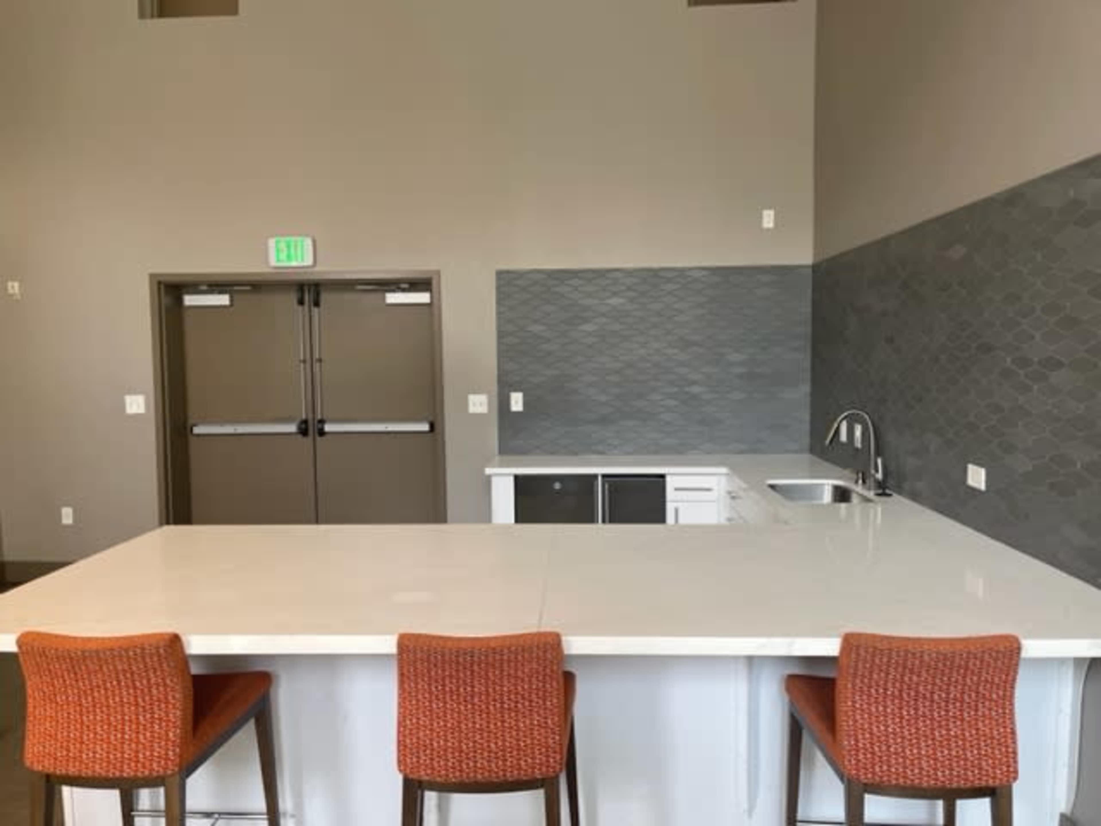 The image shows a modern kitchen with a large white countertop, three orange stools, and a gray tiled backsplash.