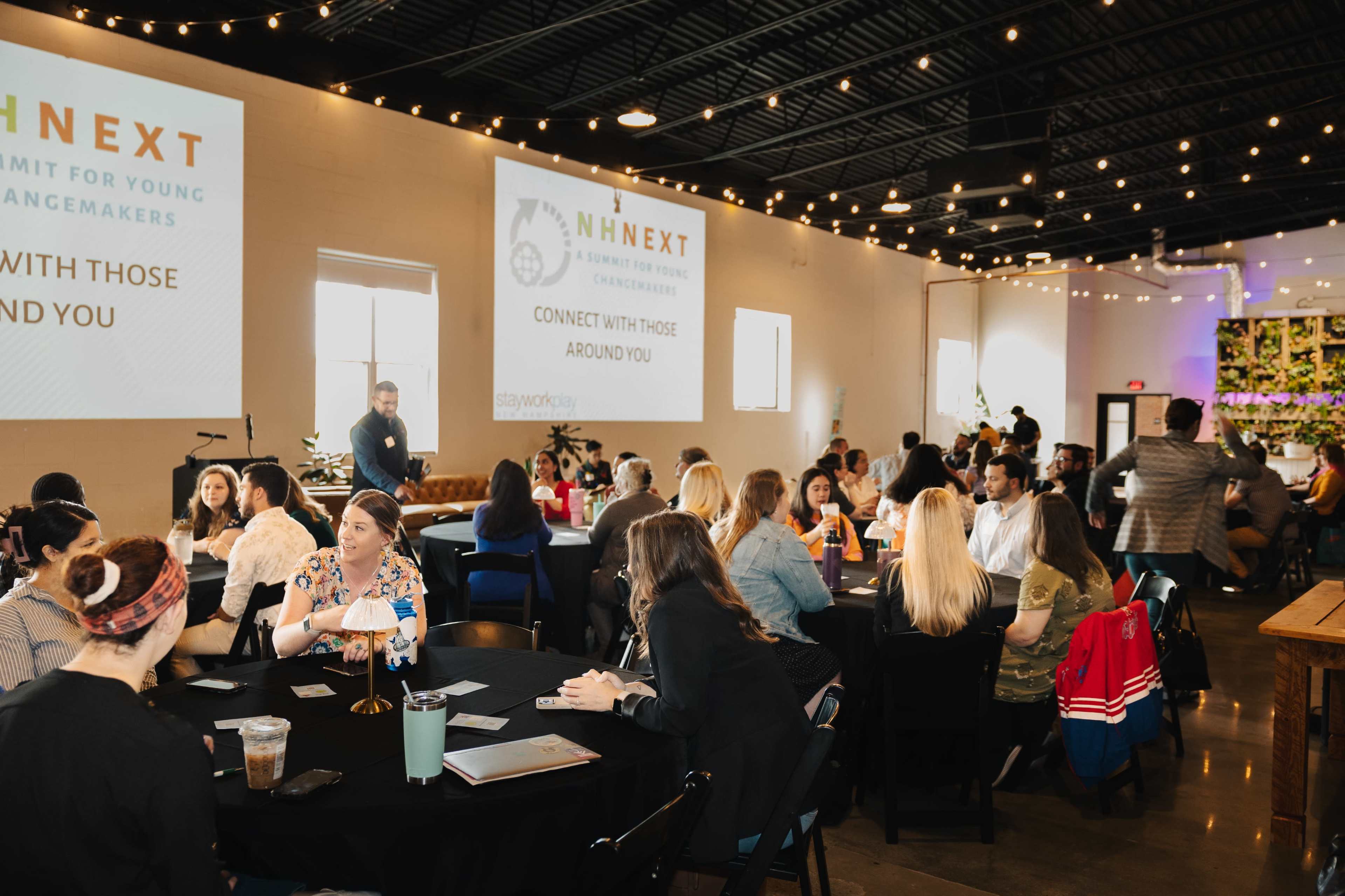 A large group of people is seated at tables in a well-lit venue, engaging in discussions during a conference marked by a presentation on a screen.