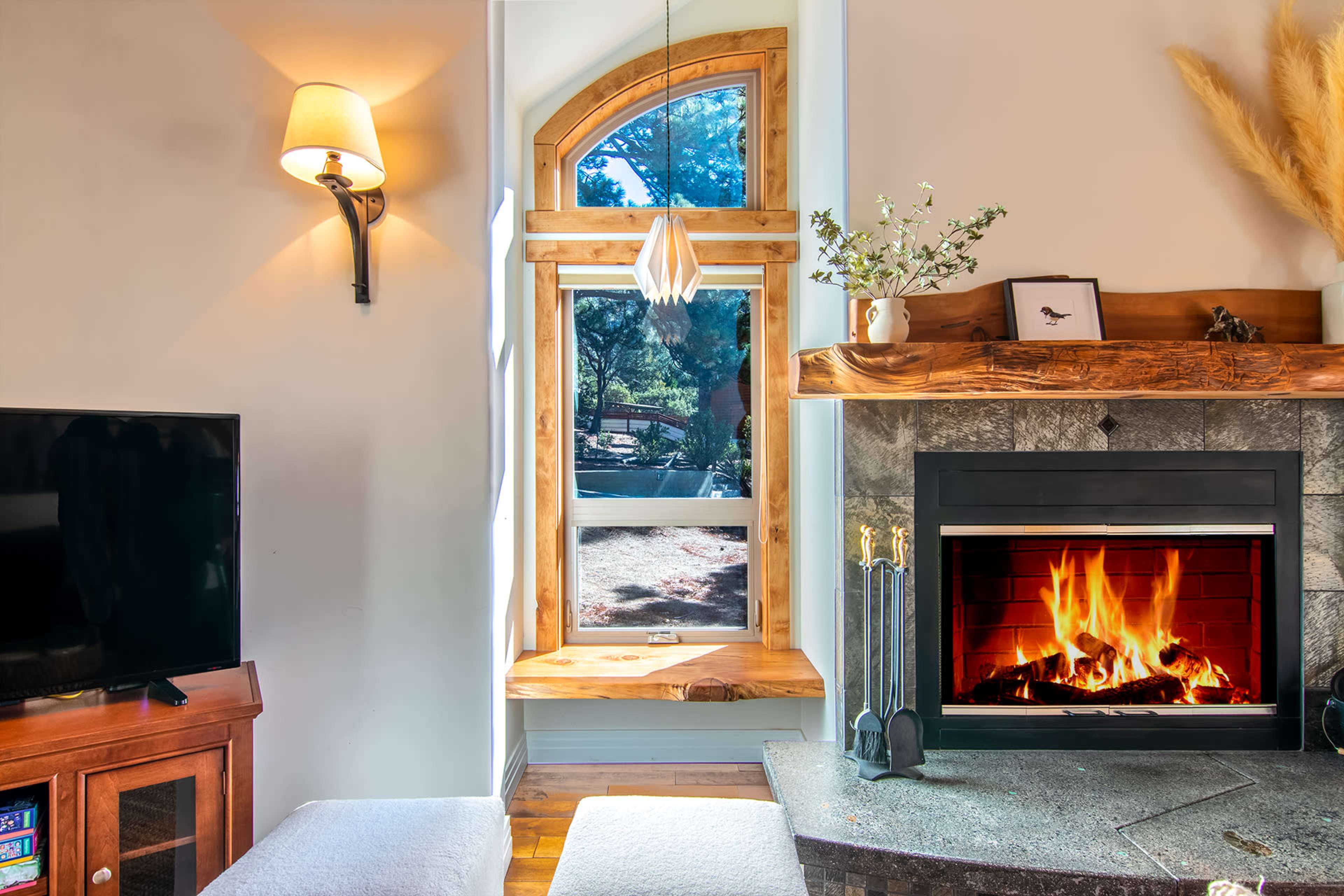 A cozy living room features a stone fireplace with a fire burning, a wooden mantle, a large window allowing natural light, and a television on a wooden cabinet.