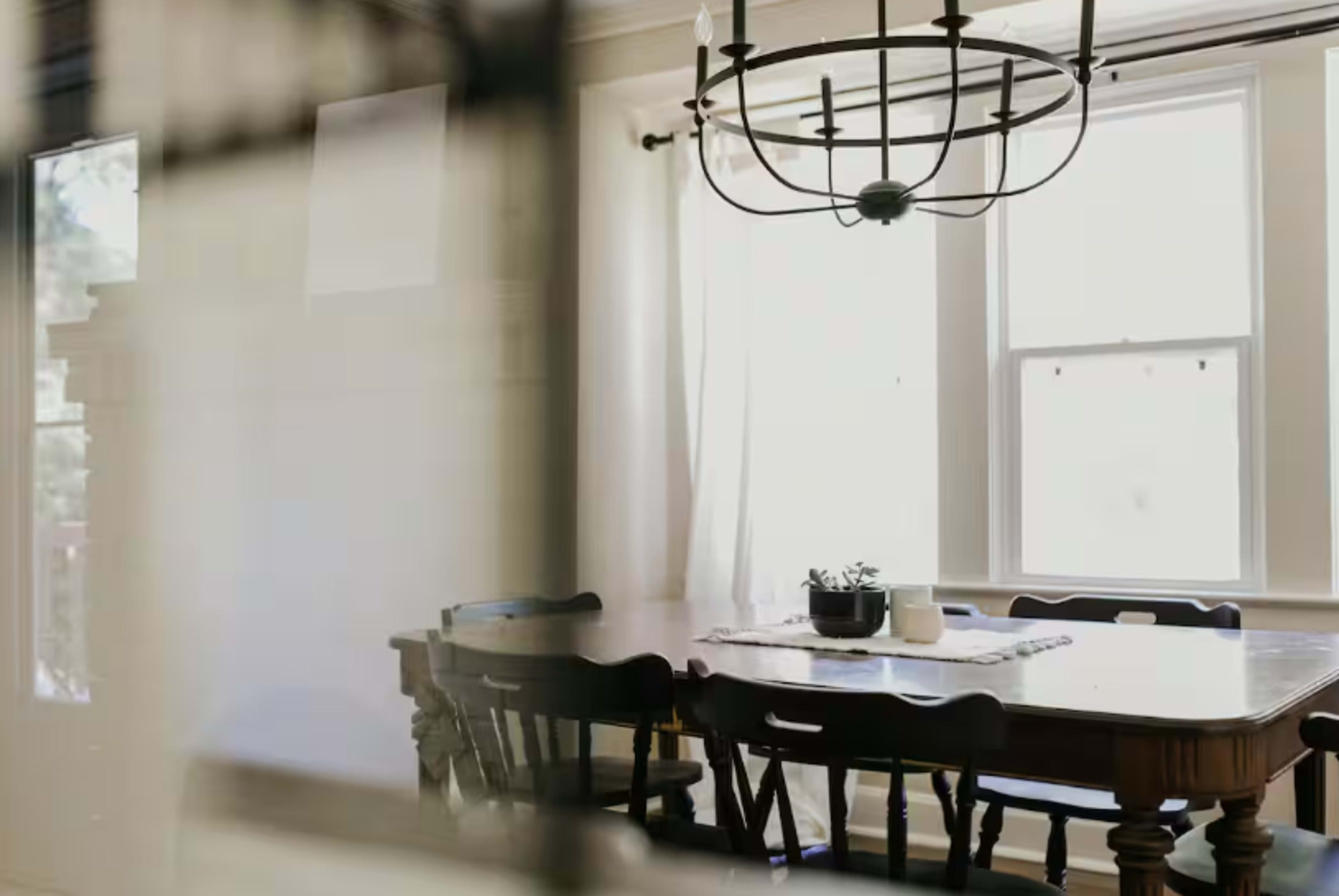 A dining room with a large wooden table, several chairs, and a chandelier, illuminated by natural light from the windows.