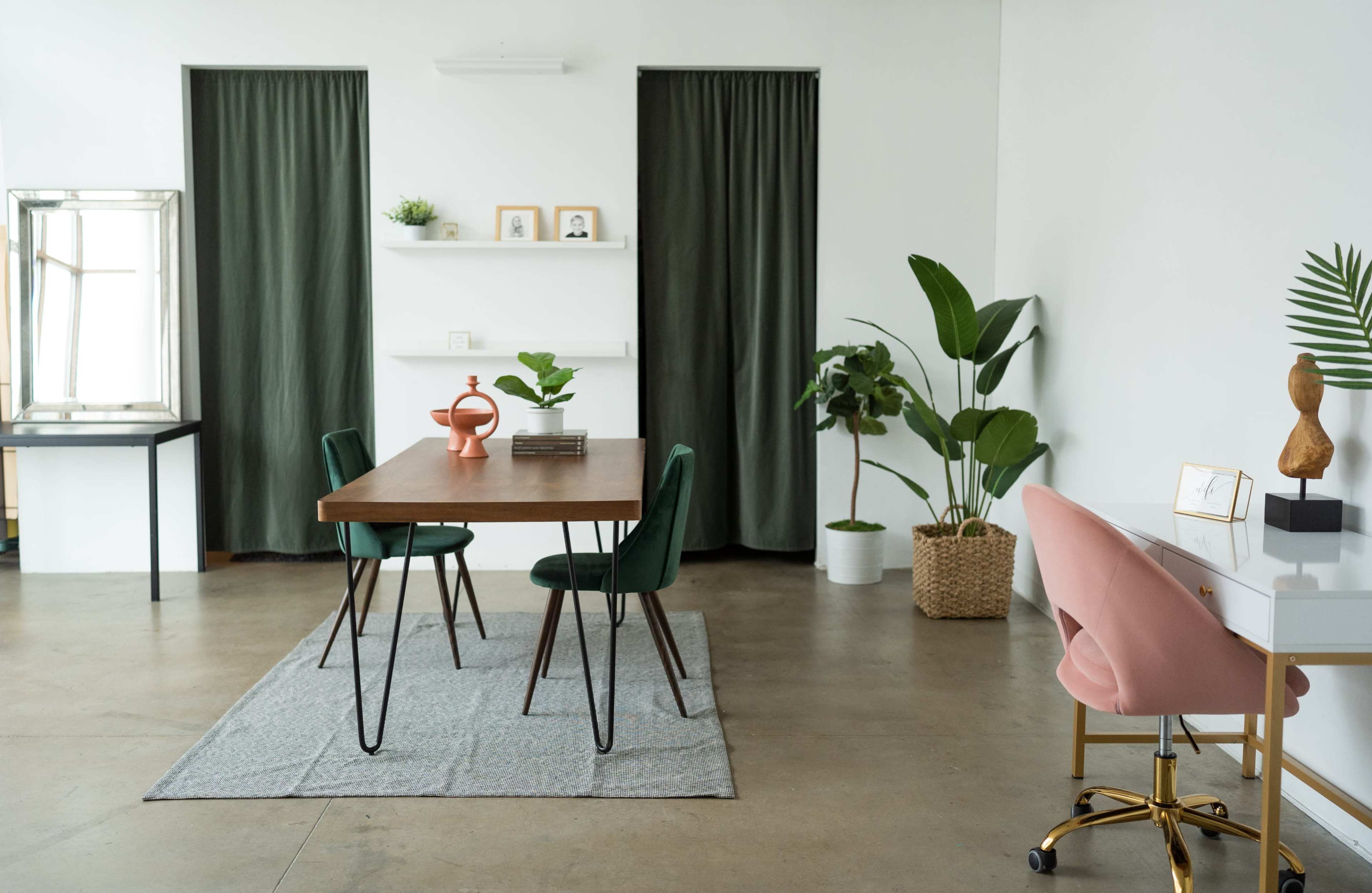 A minimalist dining area features a wooden table surrounded by green chairs, with potted plants and a desk in a bright, uncluttered space.