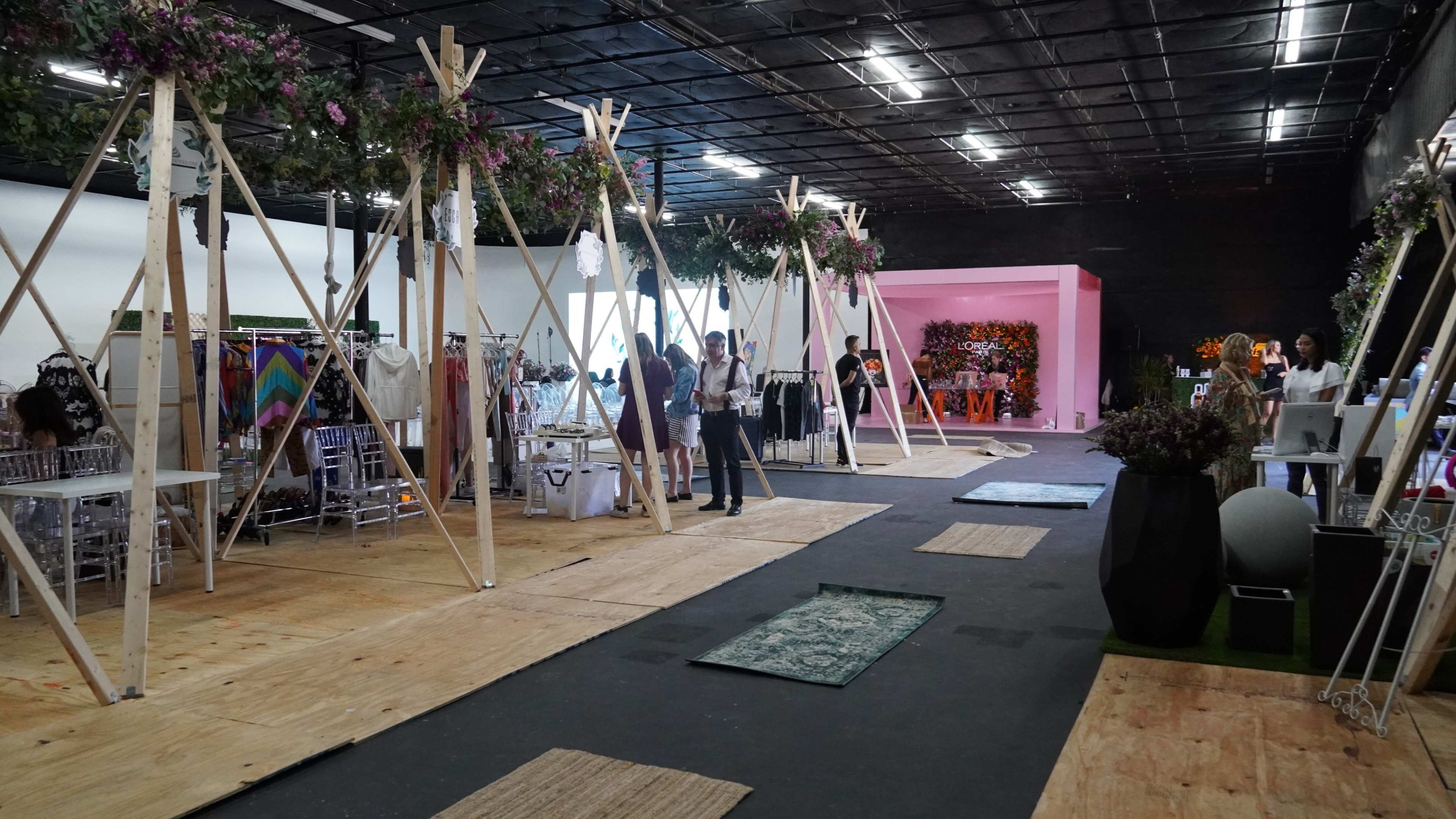 The image shows a spacious event venue with wooden structures and floral decorations, showcasing various display booths and carpeted areas for visitors.