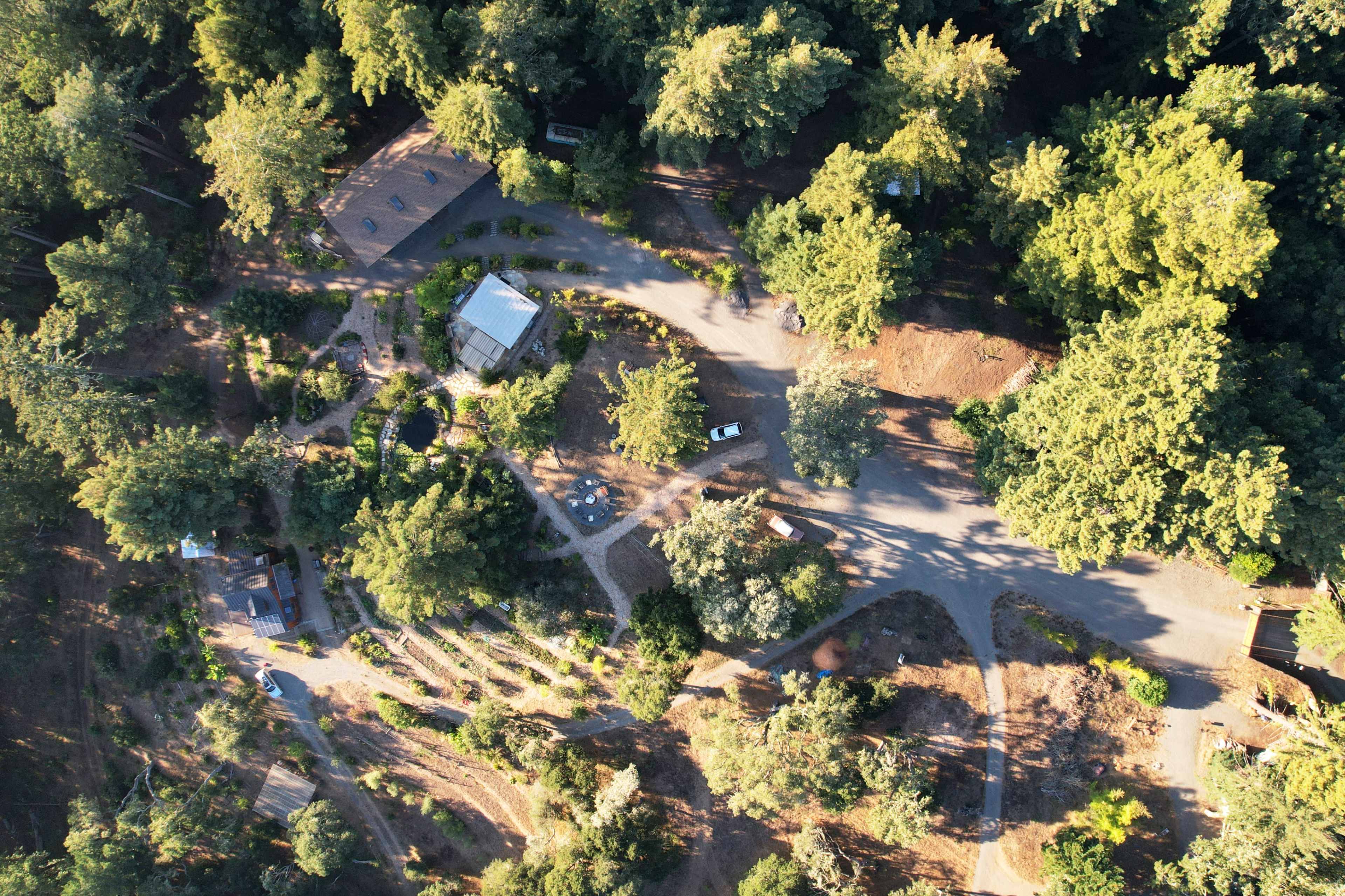 An aerial view shows a wooded area with a cluster of buildings and winding pathways amid dense trees.