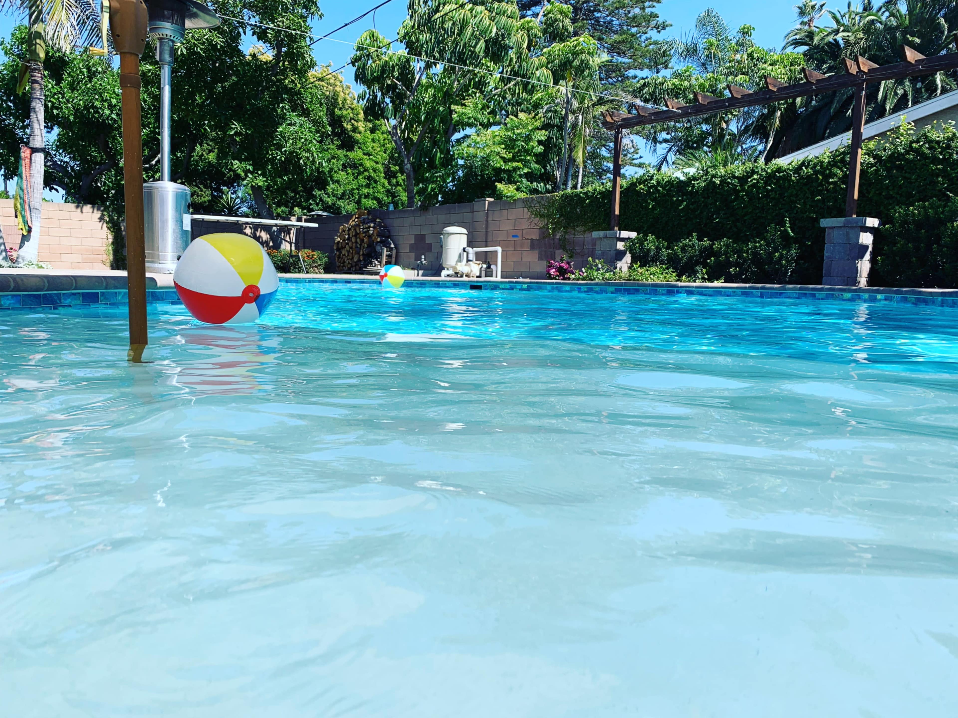 A clear swimming pool features colorful beach balls floating on its surface, surrounded by lush green trees and shrubs.