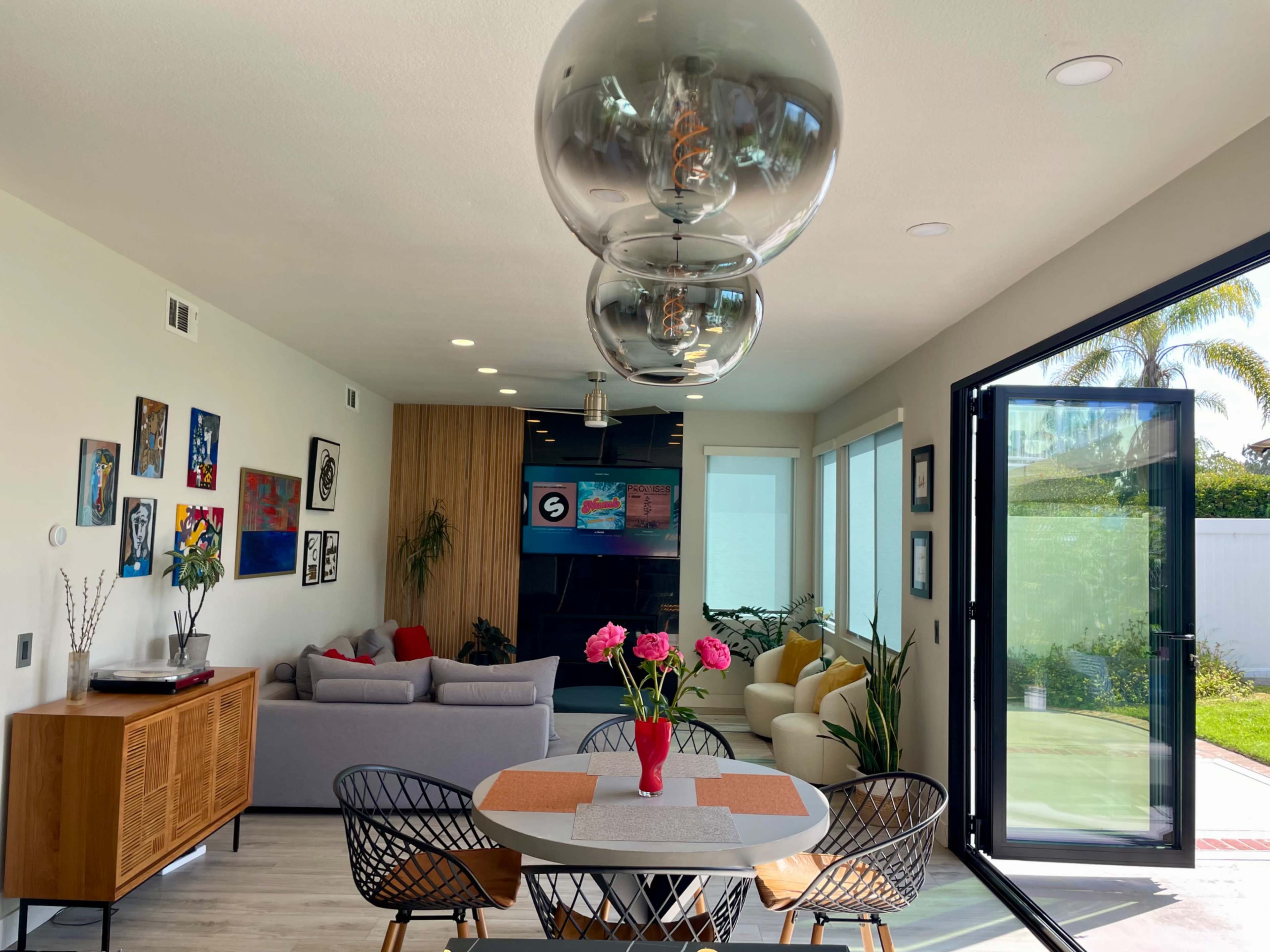 A modern living space features a dining area with a round table surrounded by black wire chairs, a sofa in the background, and large glass doors leading to a garden.