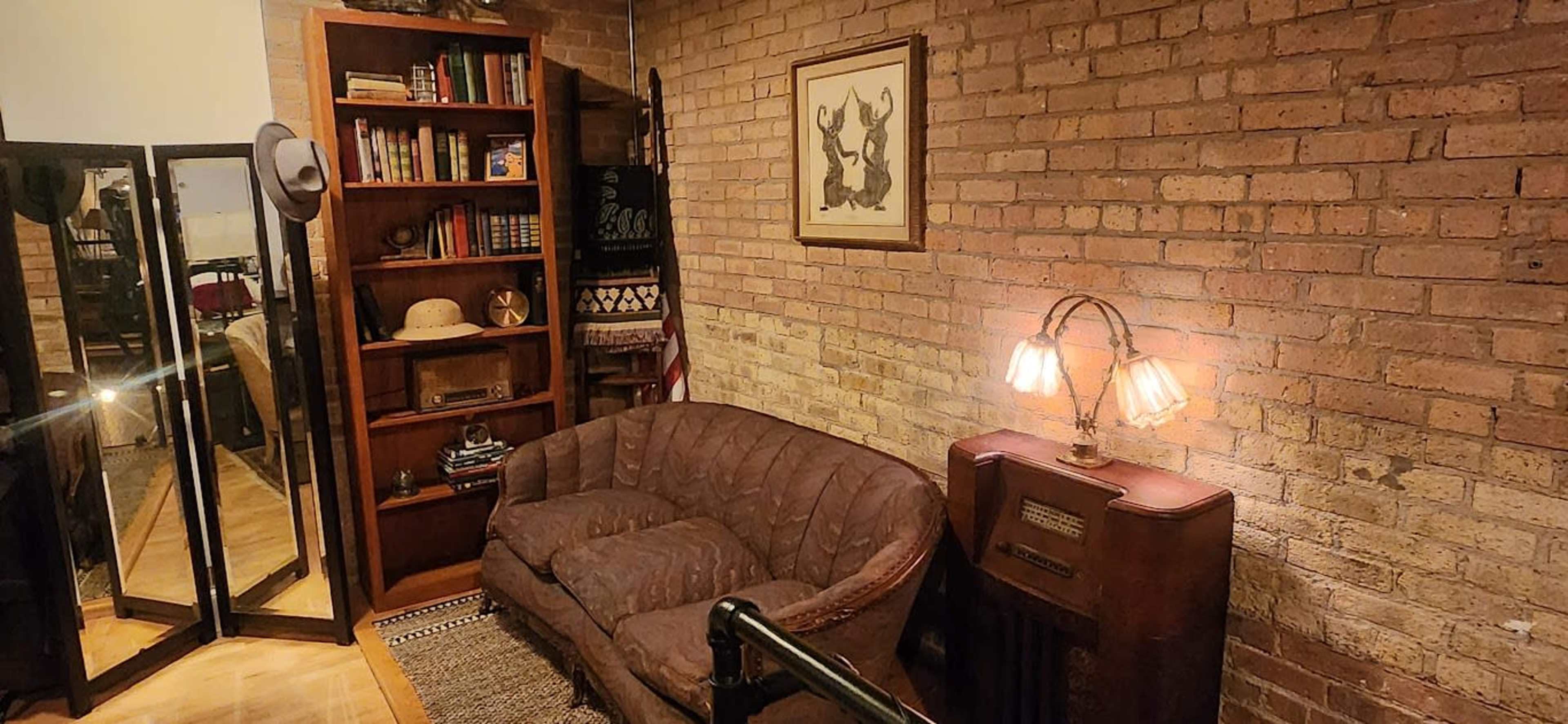 A cozy room with exposed brick walls, featuring a brown couch, a bookshelf filled with books, a vintage lamp, and a mirror reflecting the space.