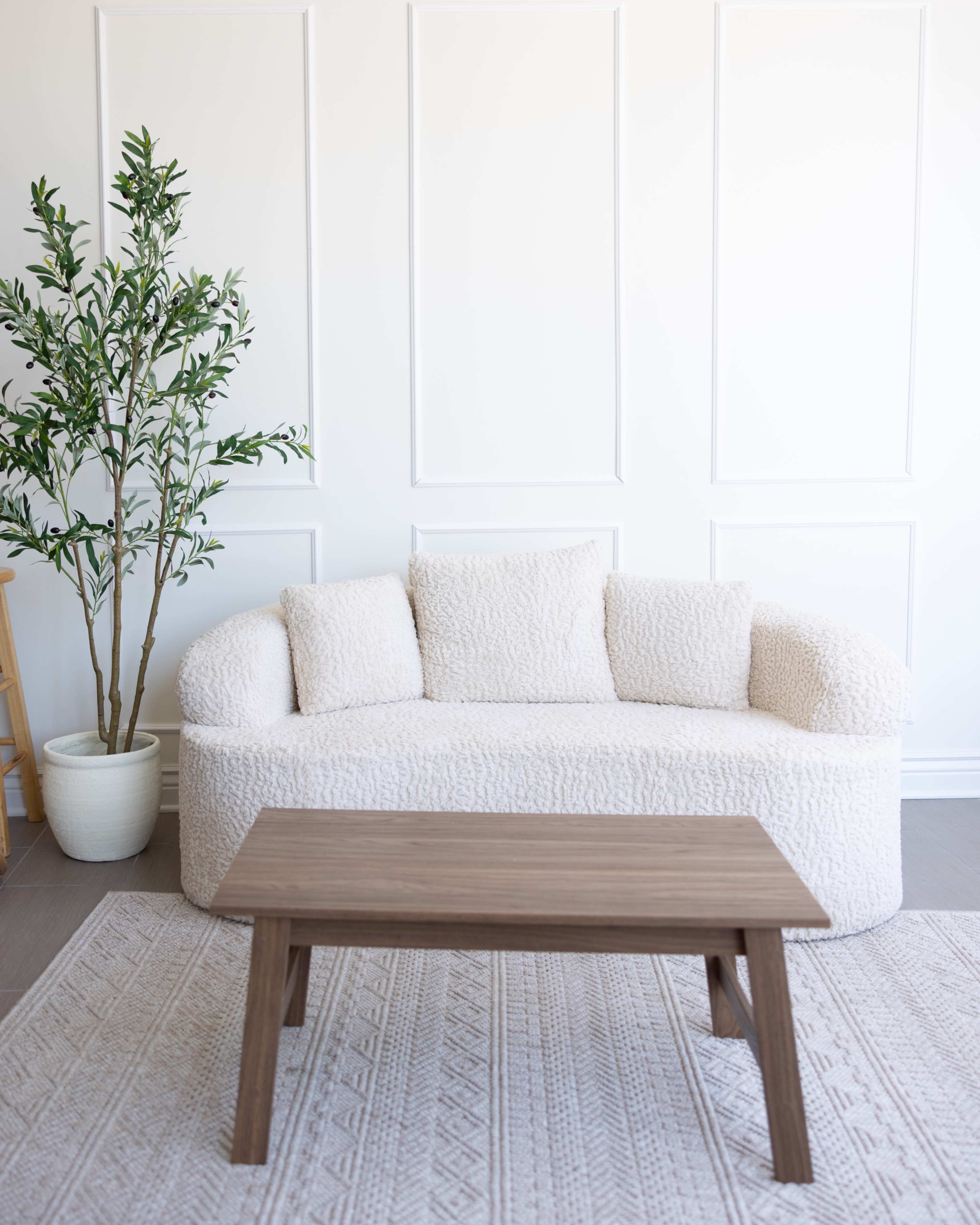 A light-colored, plush sofa with cushions sits in front of a small wooden coffee table on a patterned rug, next to a potted plant against a white paneled wall.