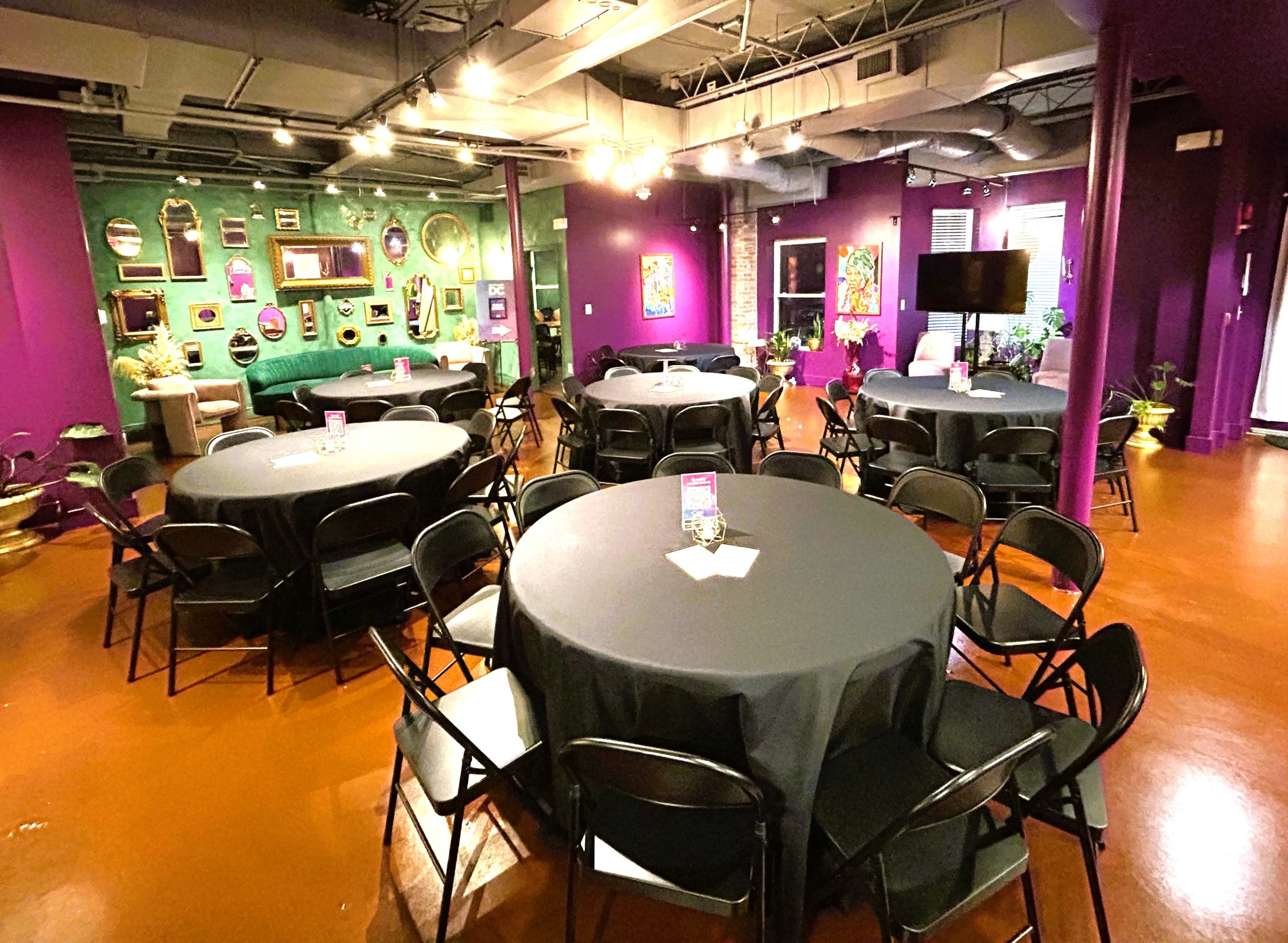A spacious event room features round tables covered with black tablecloths, surrounded by purple walls adorned with framed artwork.