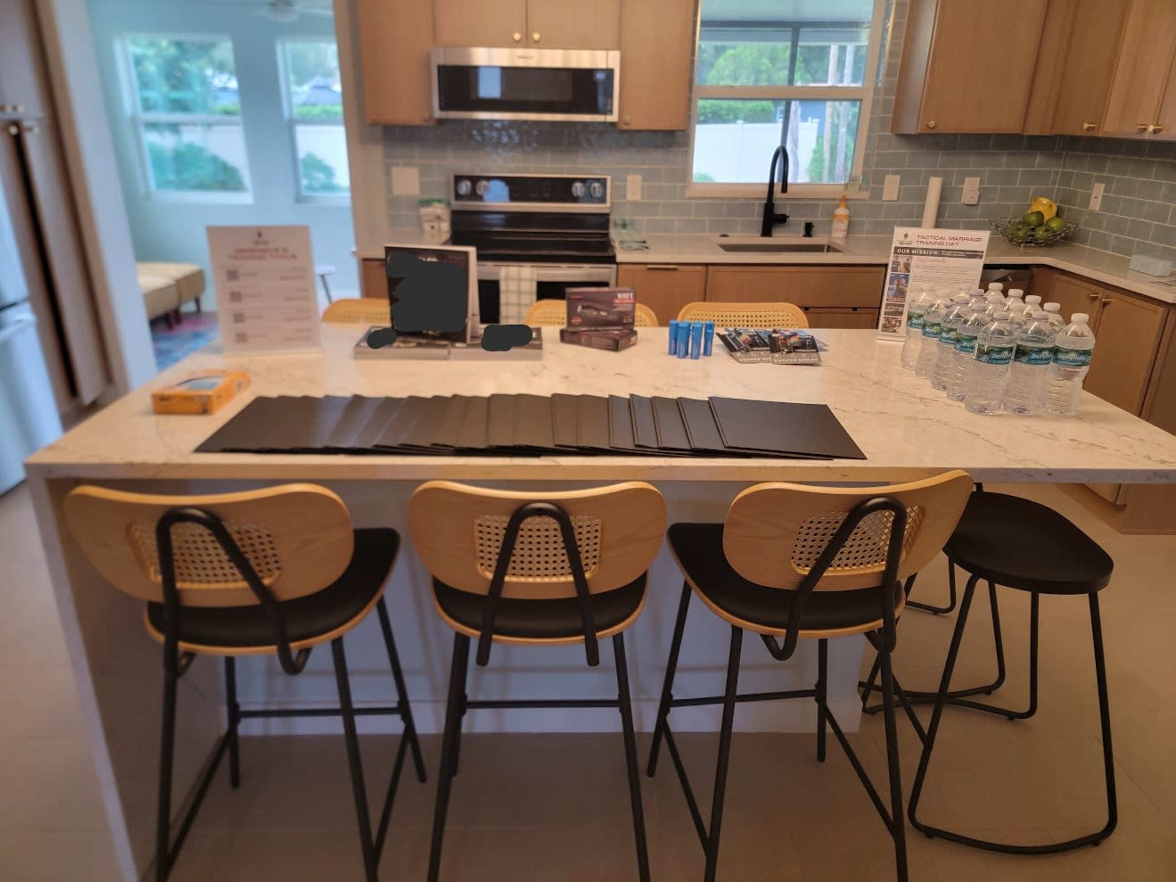A modern kitchen features a marble island with seating, water bottles, and various materials laid out for a presentation.