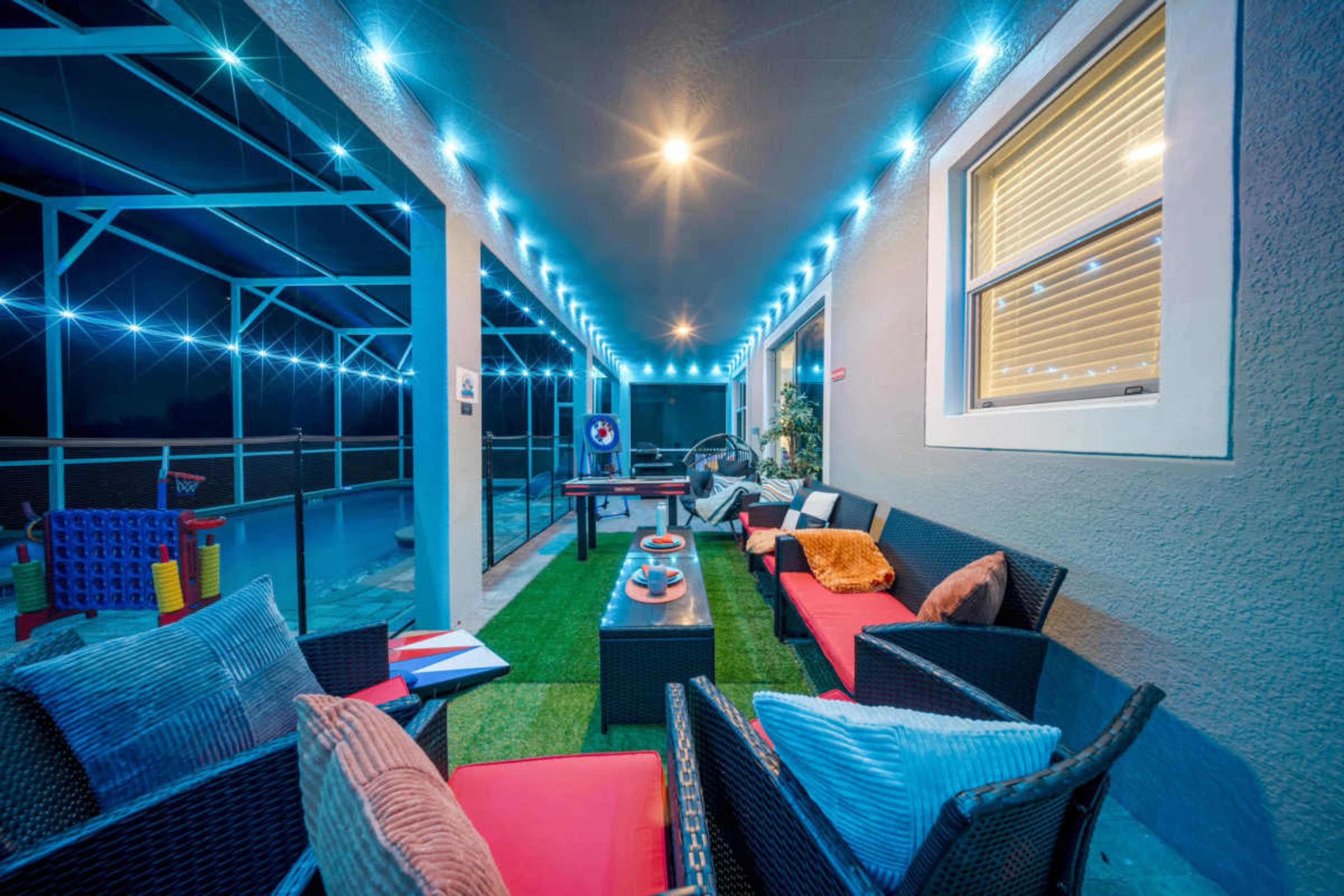The image depicts a modern outdoor patio area with comfortable seating, decorative lighting, and a view of a swimming pool.