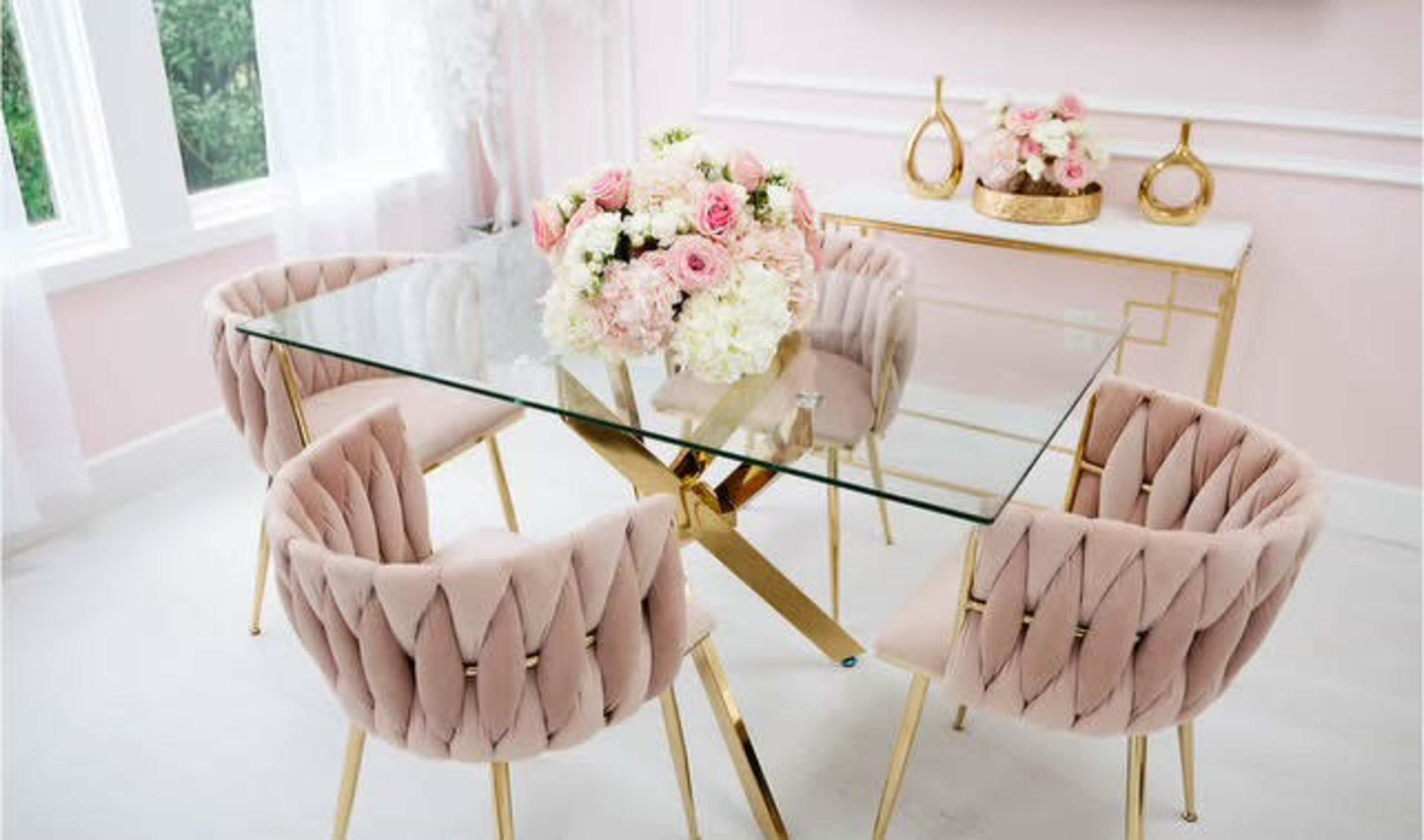 A glass dining table surrounded by four pink upholstered chairs is set in a room with light pink walls and decorative floral arrangements.
