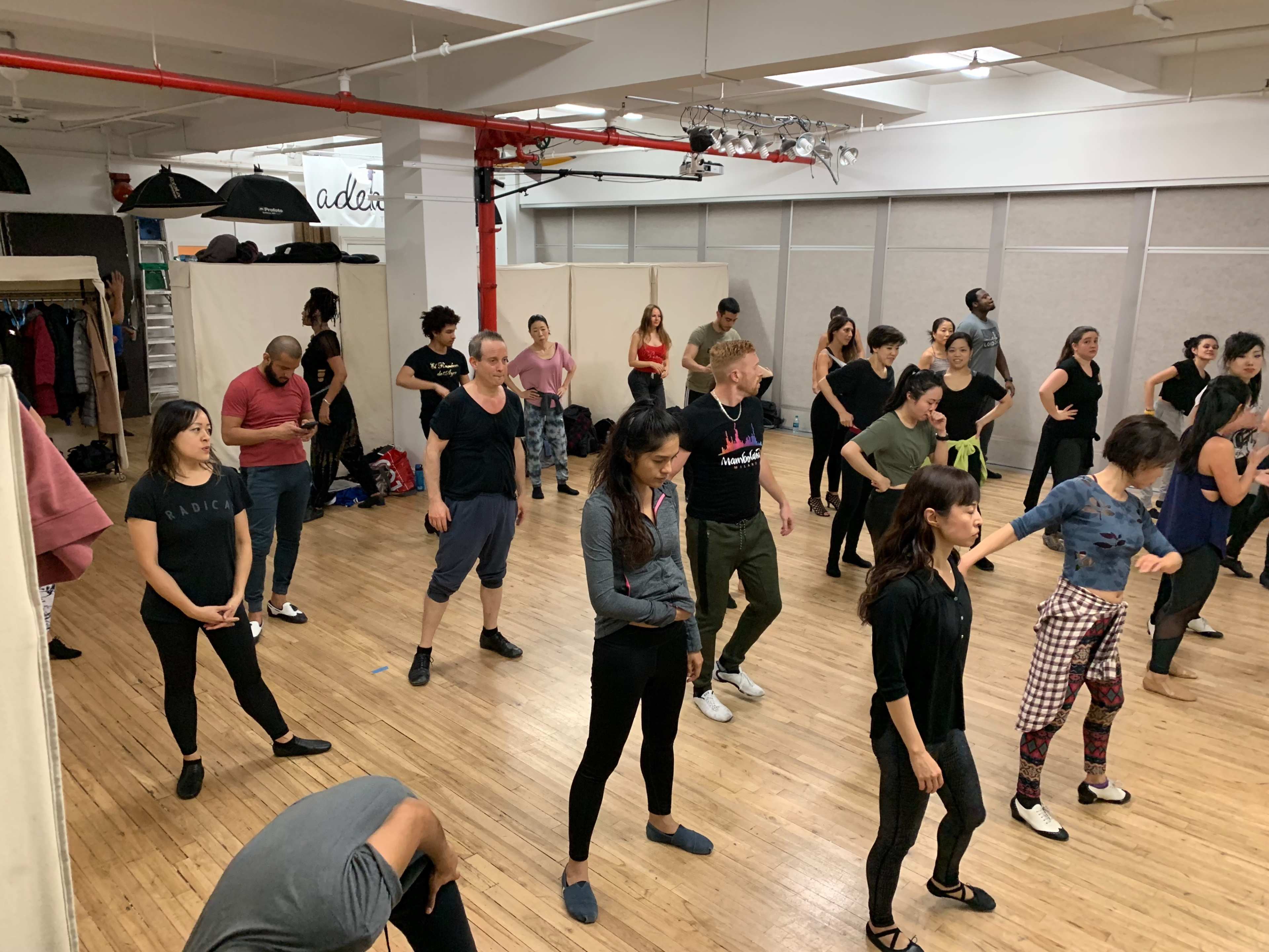 A large group of people is practicing dance in a spacious studio with wooden flooring and red pipes.