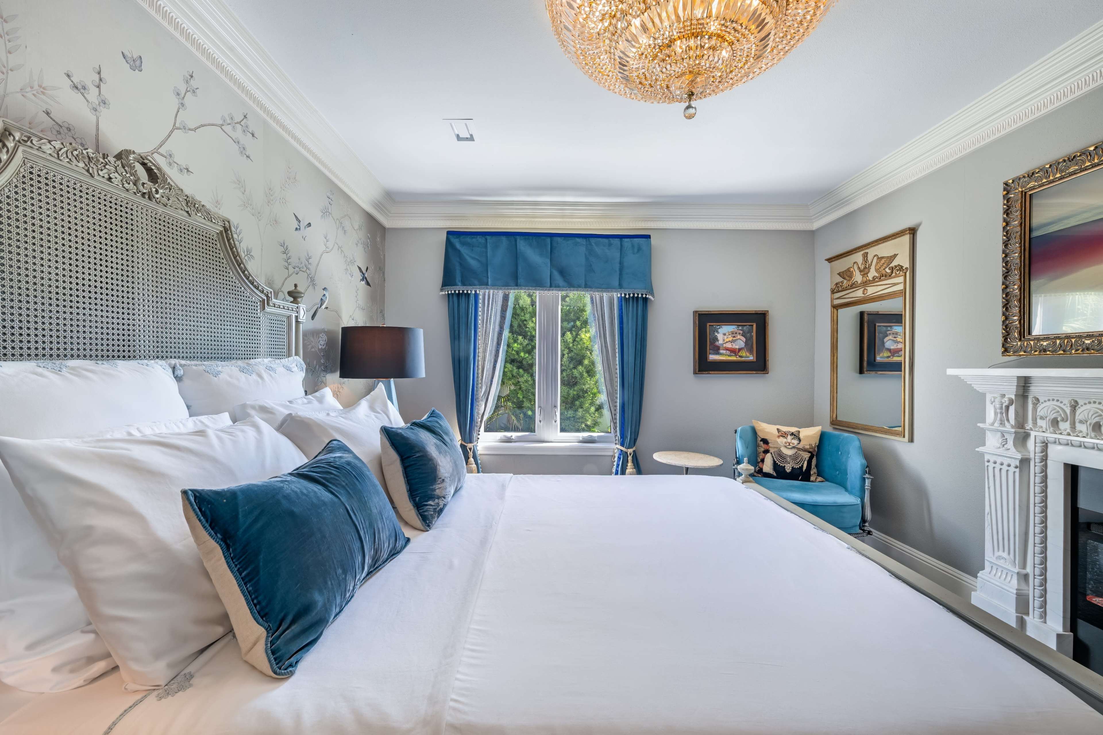 The image shows a neatly arranged bedroom featuring a large bed with decorative pillows, a blue upholstered chair, a window with blue curtains, and a fireplace with a mirror above it.