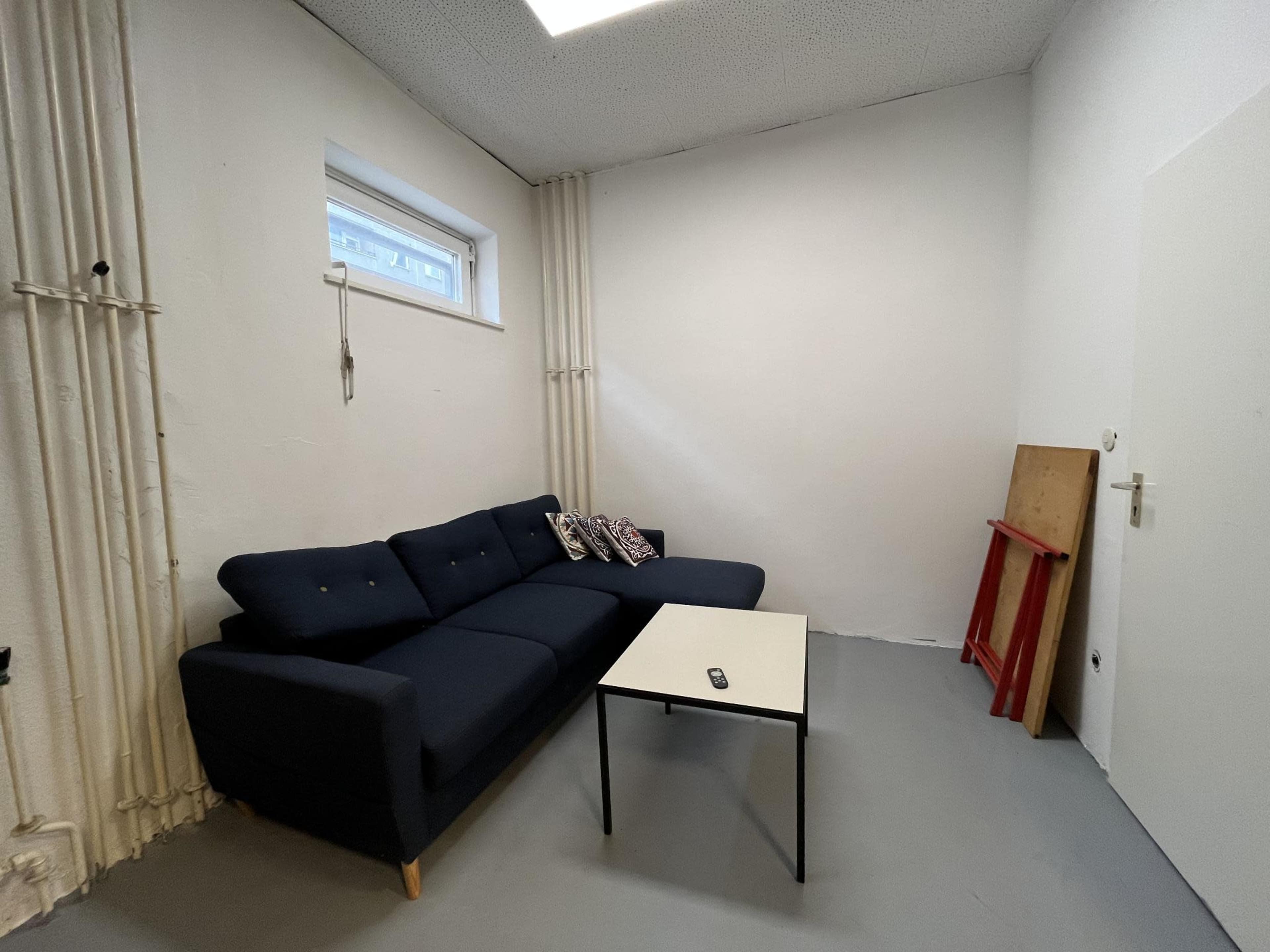A small room features a dark sofa with cushions and a white coffee table in front, along with a red board leaning against the wall.