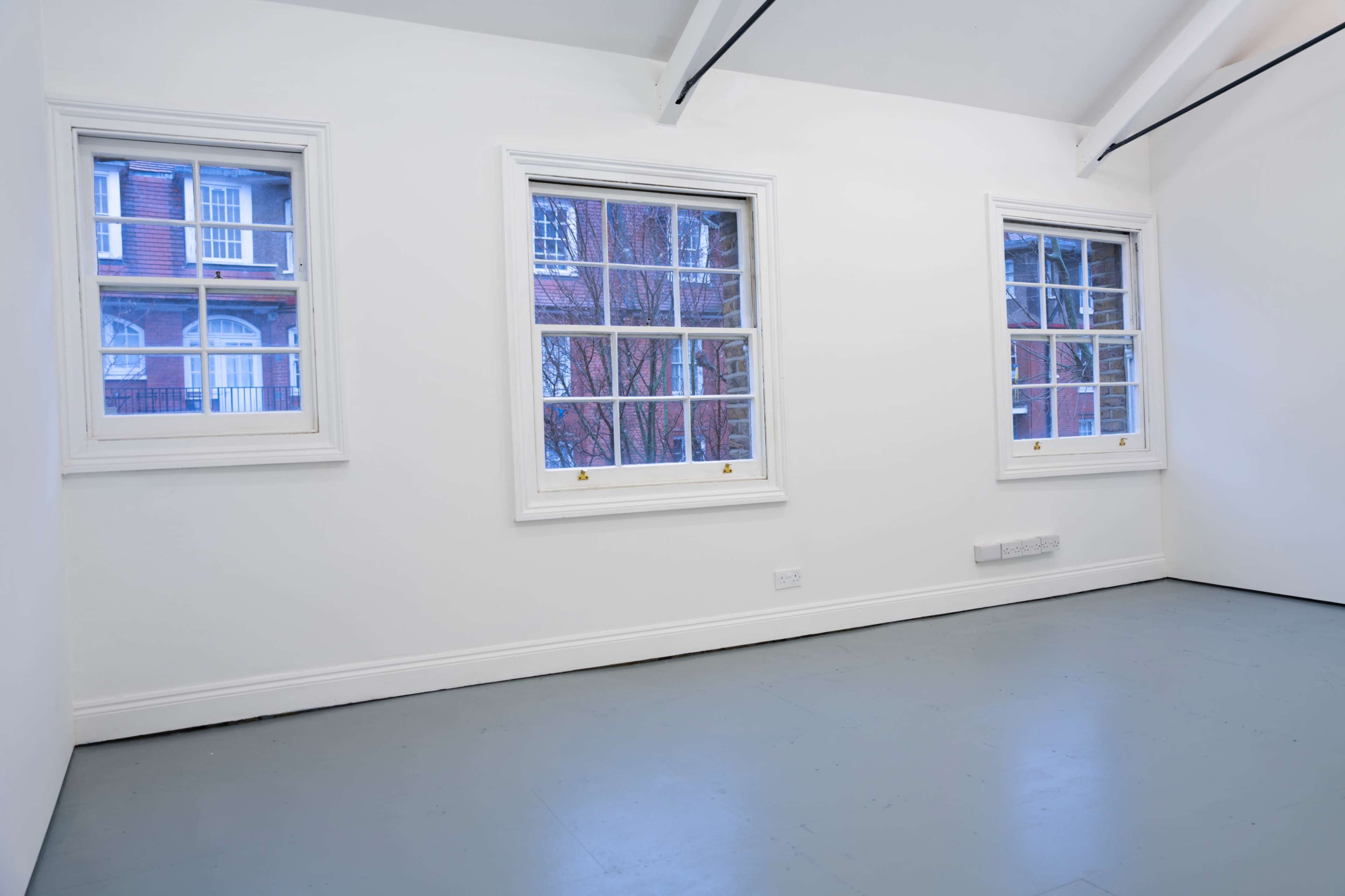 The image shows a bright, empty room with three white-framed windows revealing a view of an exterior brick wall.