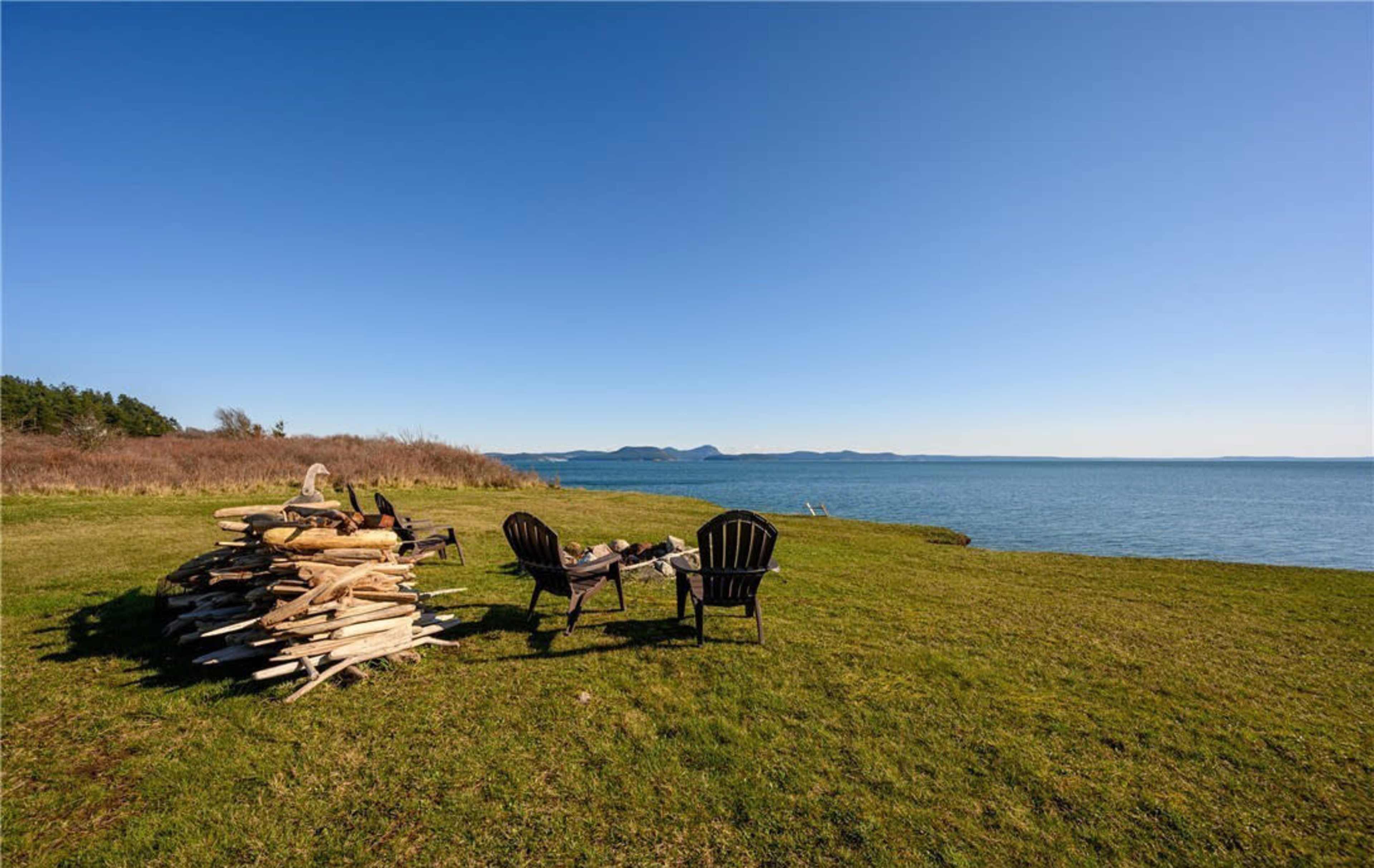 Two Adirondack chairs sit facing the water beside a neatly stacked pile of firewood on a grassy shoreline.