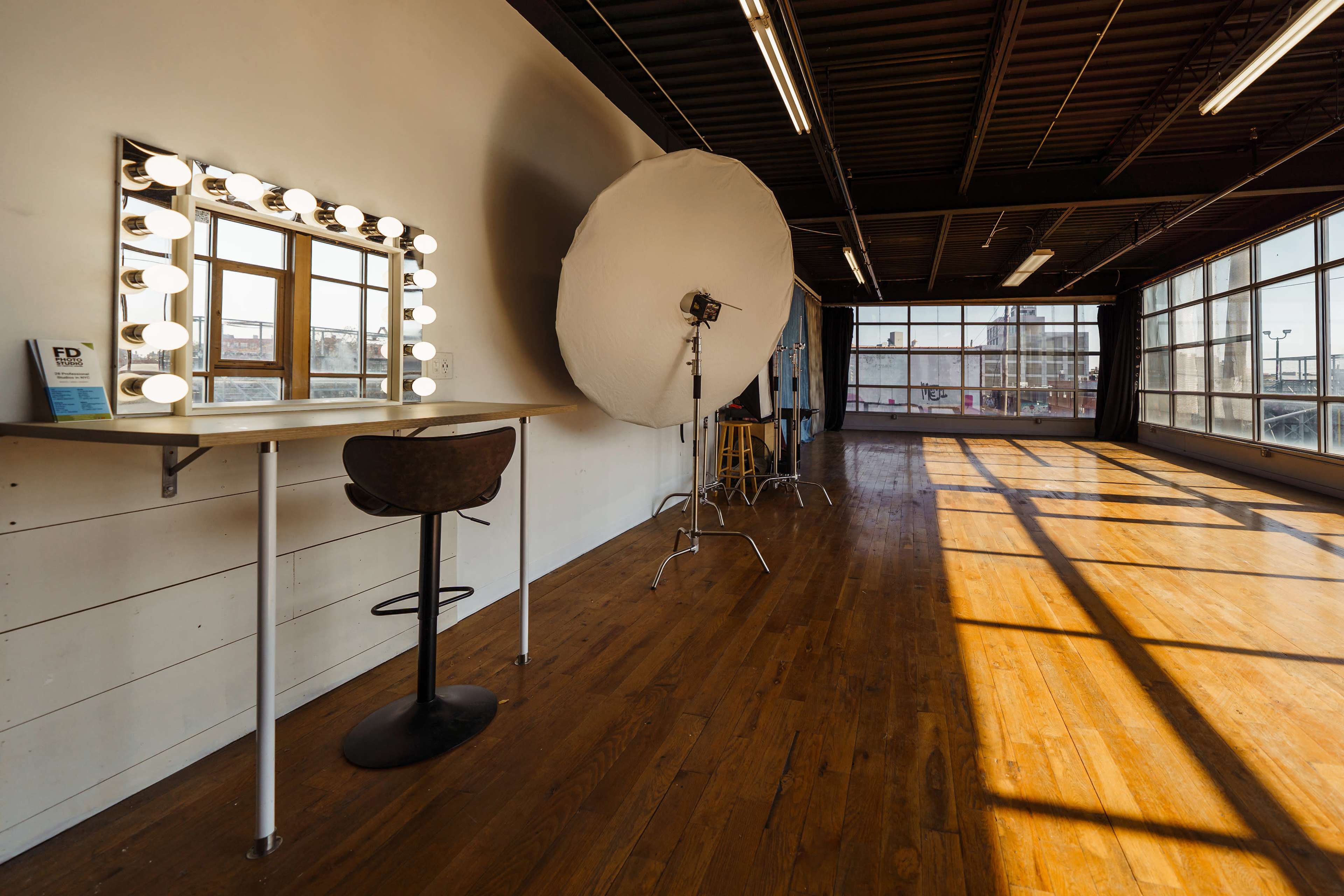 A photography studio with a mirror surrounded by lights, a softbox, and wooden flooring.
